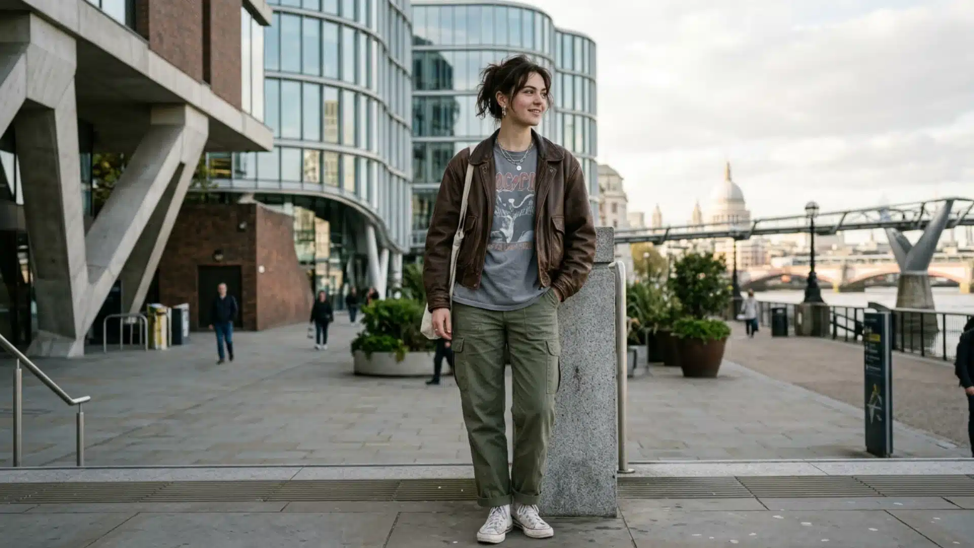 A woman in a leather jacket, graphic tee, and green cargo pants stands outdoors by a city river walk, looking aside