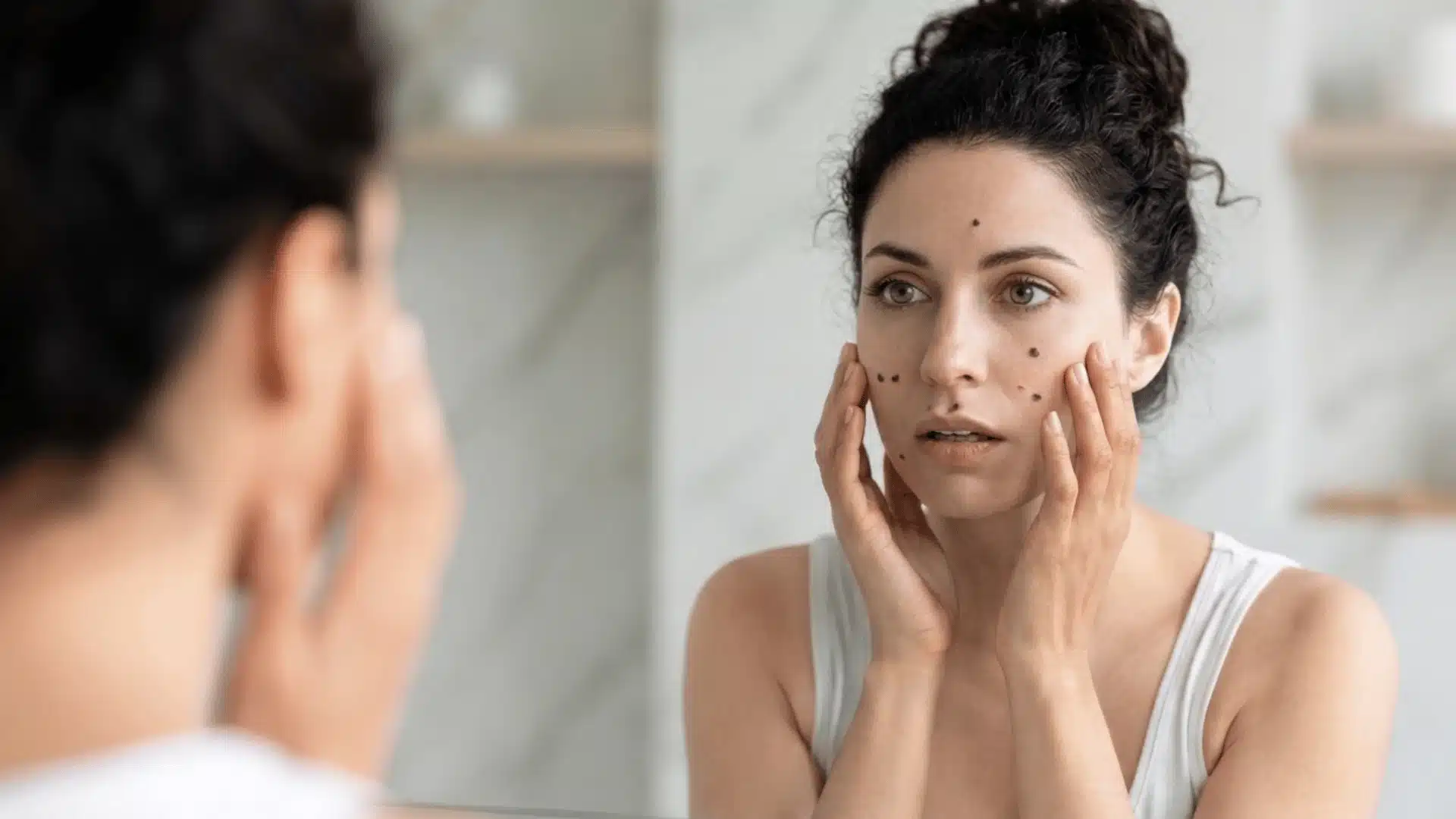 A woman with dark, curly hair tied up looks anxiously into a bathroom mirror, touching her face to examine skin moles
