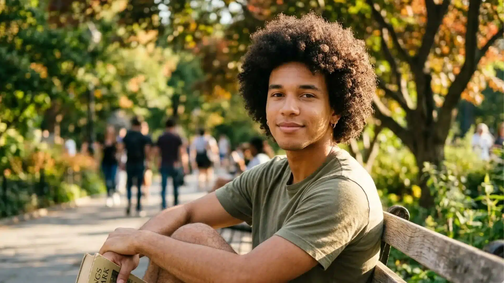 A young man with an afro sitting on a park bench, holding a book with leafy trees in the background.