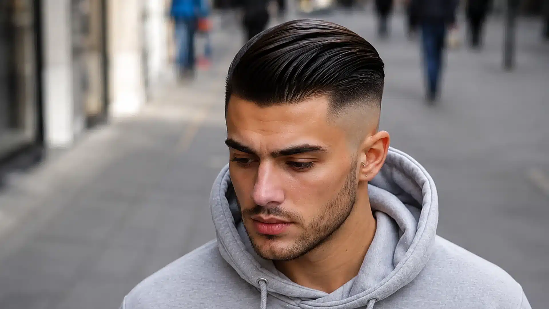 A young man with slicked-back hair, a high fade haircut, and short stubble, wearing a grey hoodie on a city street
