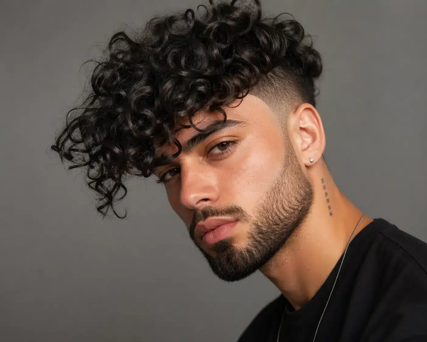 A young man with voluminous dark curls and a sharp skin fade poses against a grey background. (88 characters)
