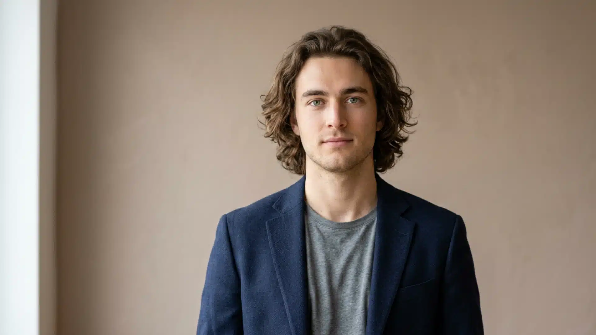 A young man with wavy brown hair, wearing a grey t-shirt and a navy blue blazer, posing against a beige wall..009