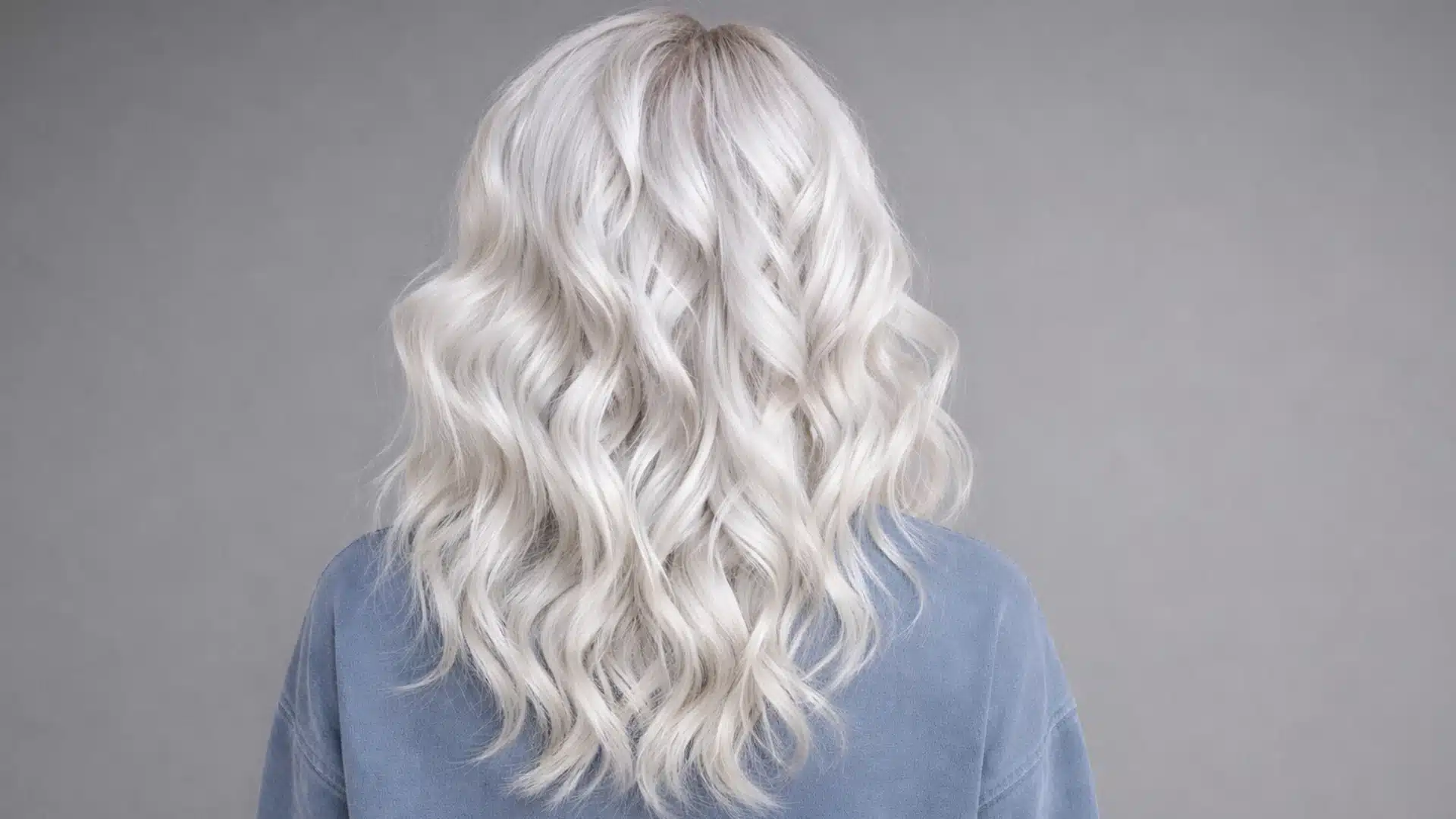 Back view of a woman with long icy silver blonde wavy hair with soft texture and volume, against a solid dark grey background with focus on hair color and shine