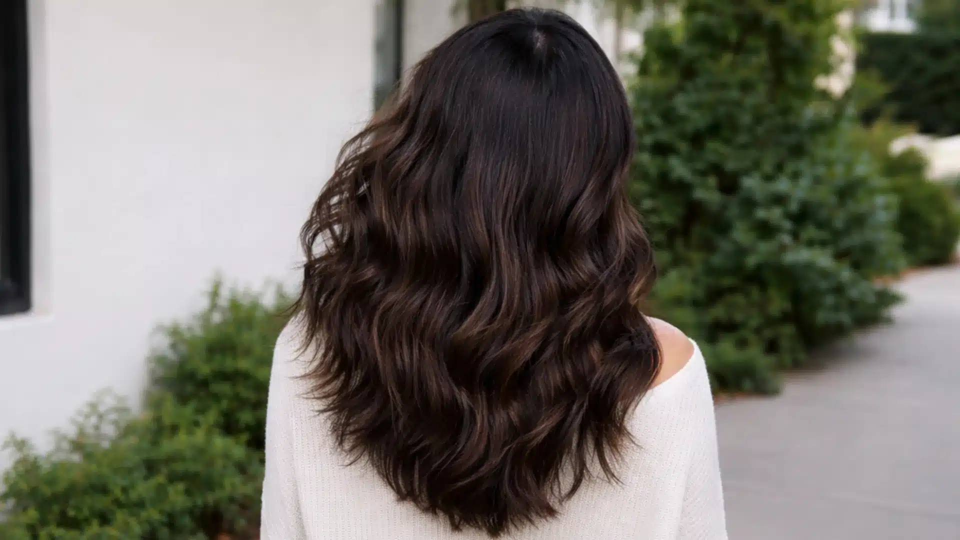 Back view of woman with long wavy dark brown hair standing outdoors, with more background visible in a soft natural setting