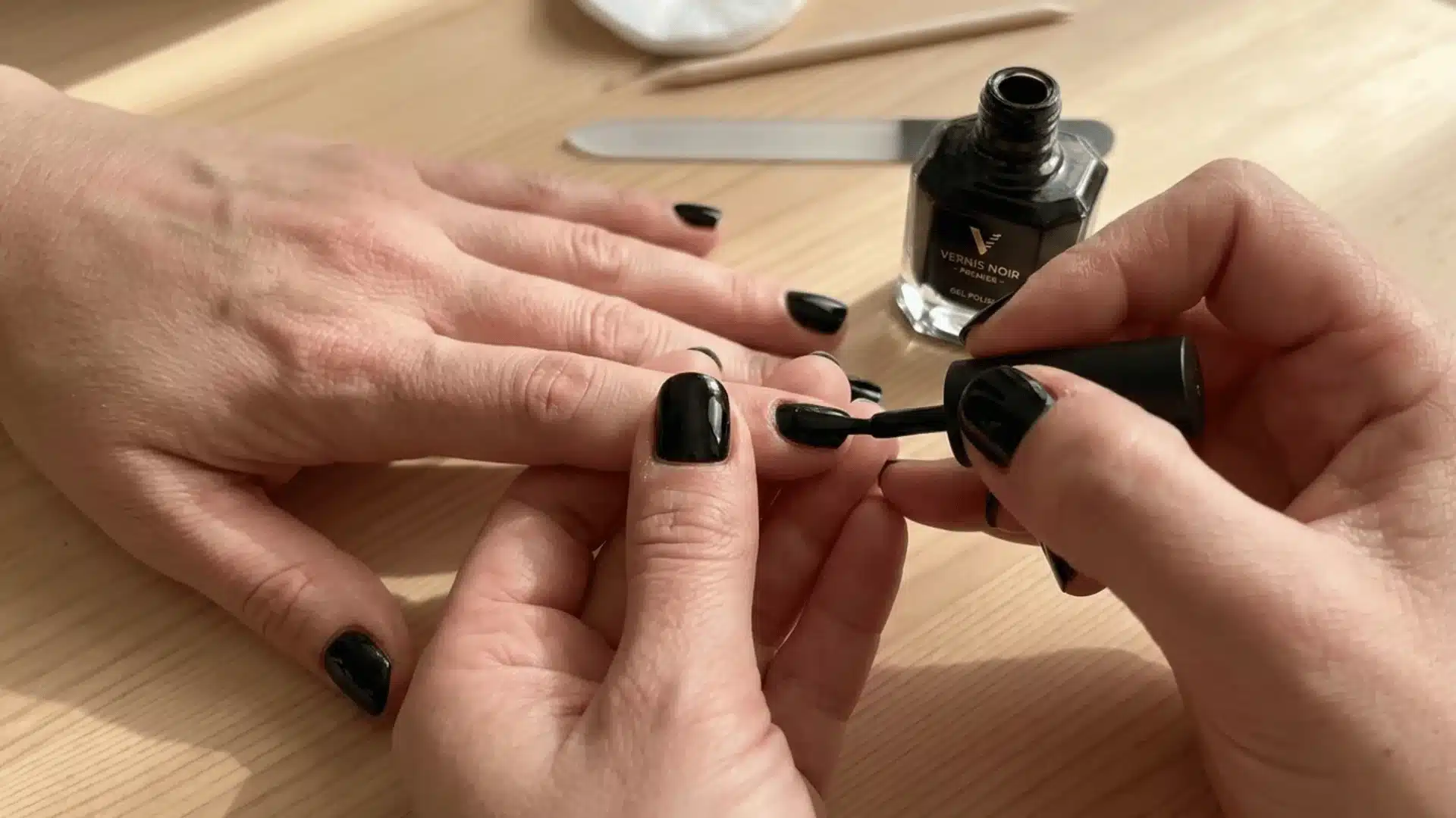 Close-up of black gel nail polish being applied to a fingernail on a wooden table with manicure tools nearby