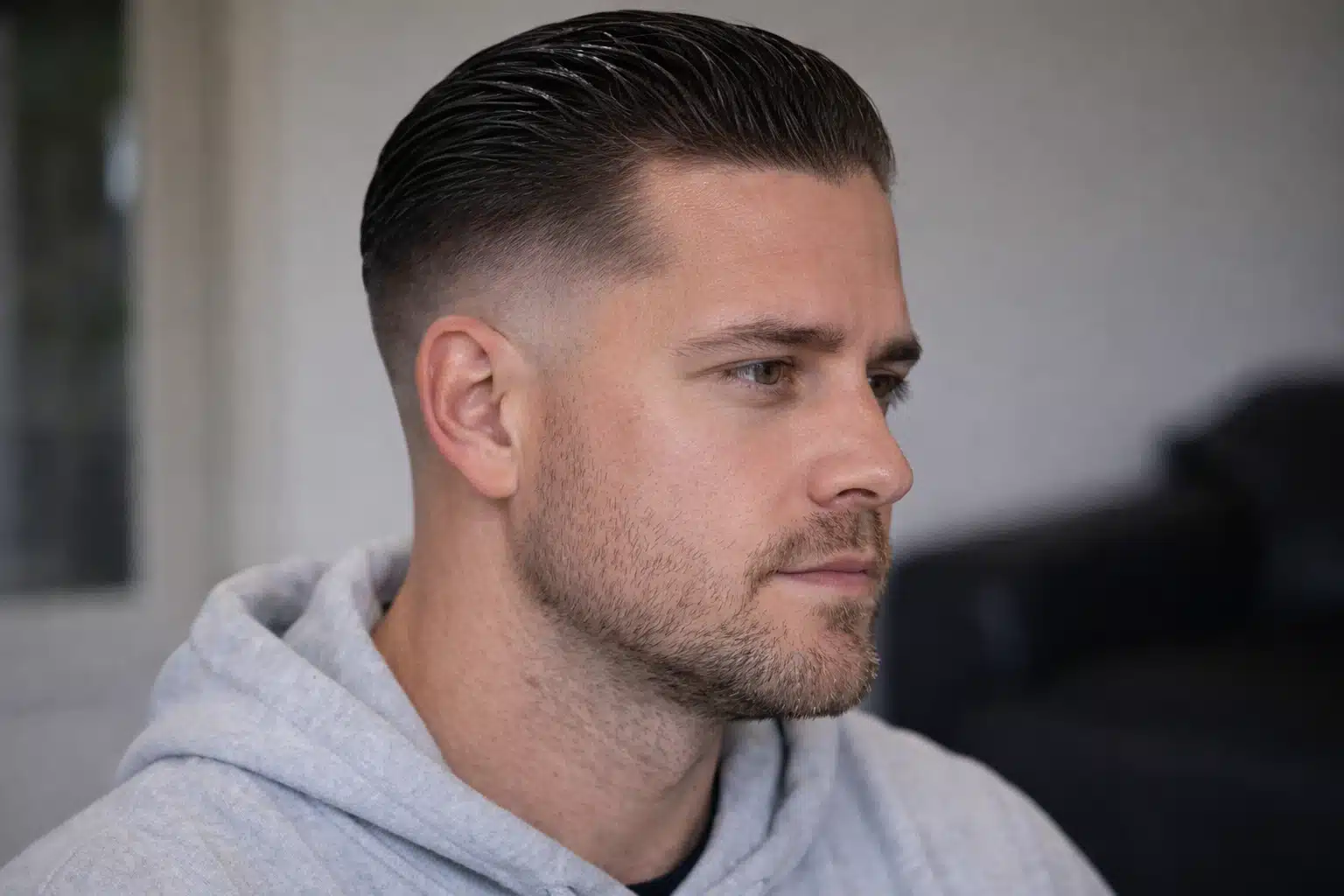 Close-up of man with wet look high fade slick back haircut, light stubble, wearing gray hoodie indoors with soft lighting lit