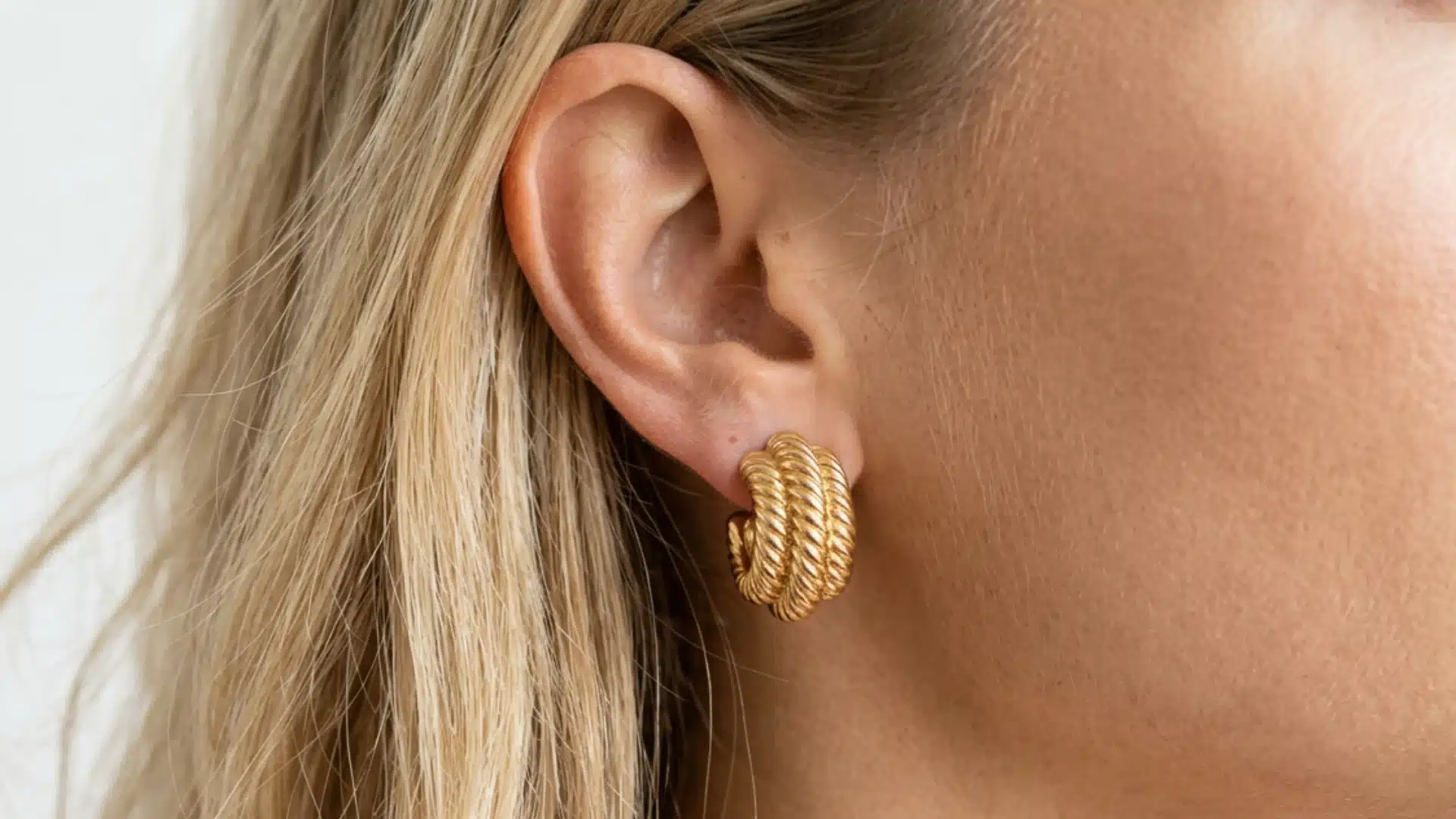 Close-up of a blonde woman's ear, wearing a chunky, ribbed gold j-hoop earring with a twisted rope design