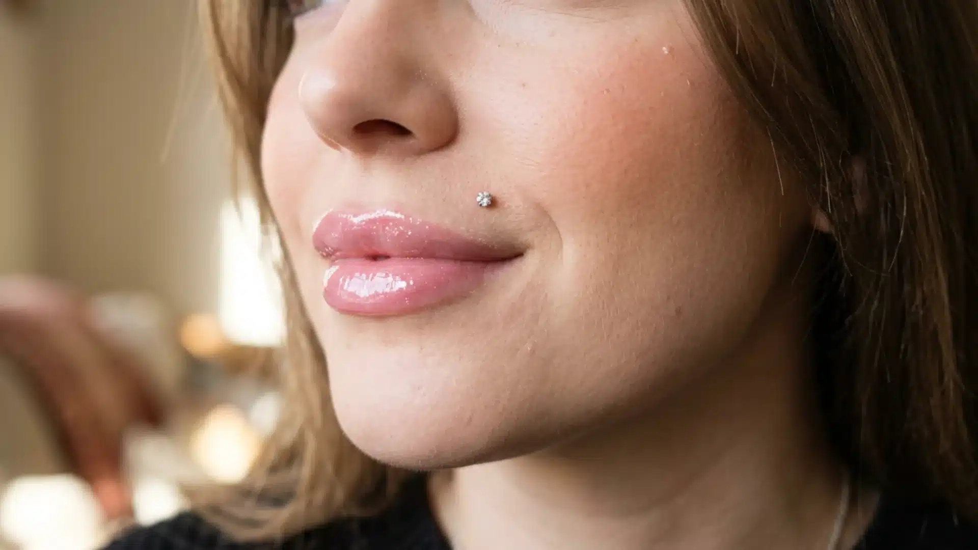 Close-up of a person smiling with glossy pink lips and a sparkling Monroe piercing above their upper lip