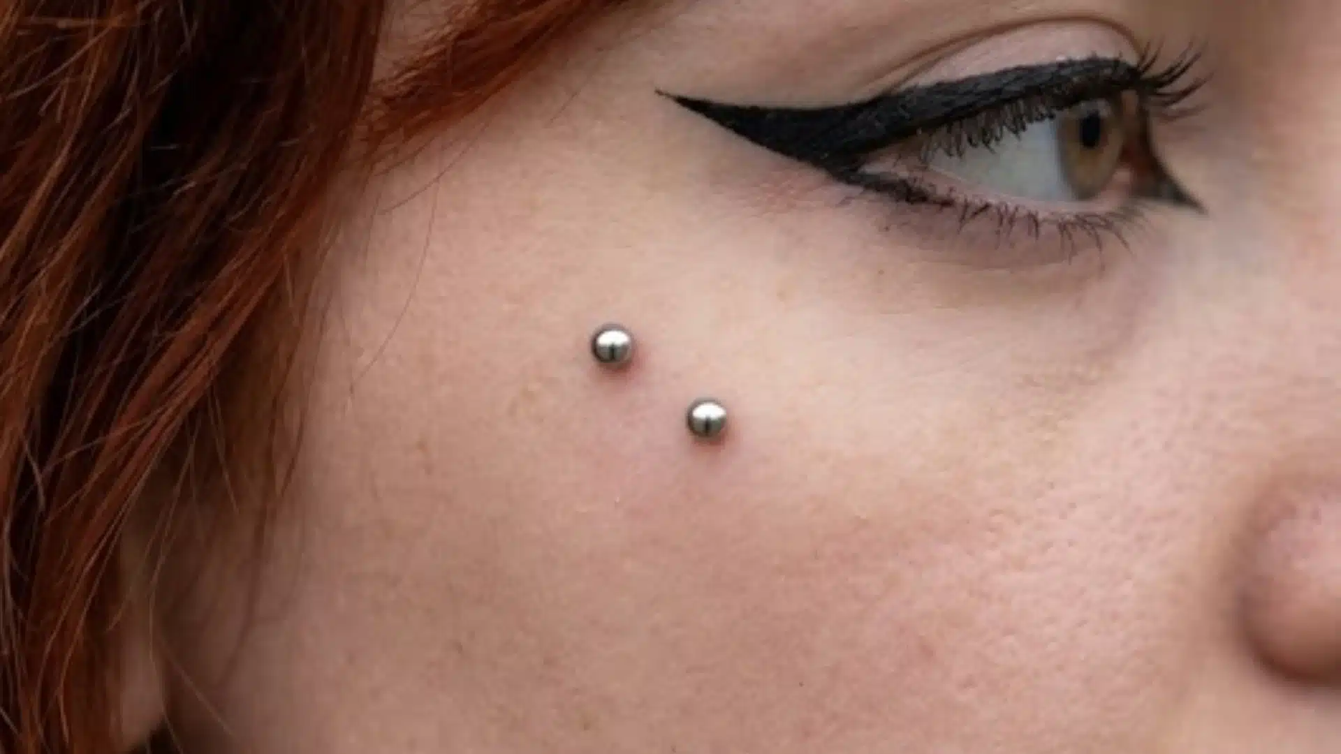 Close-up of a person with winged eyeliner and a double dermal surface piercing on their cheek
