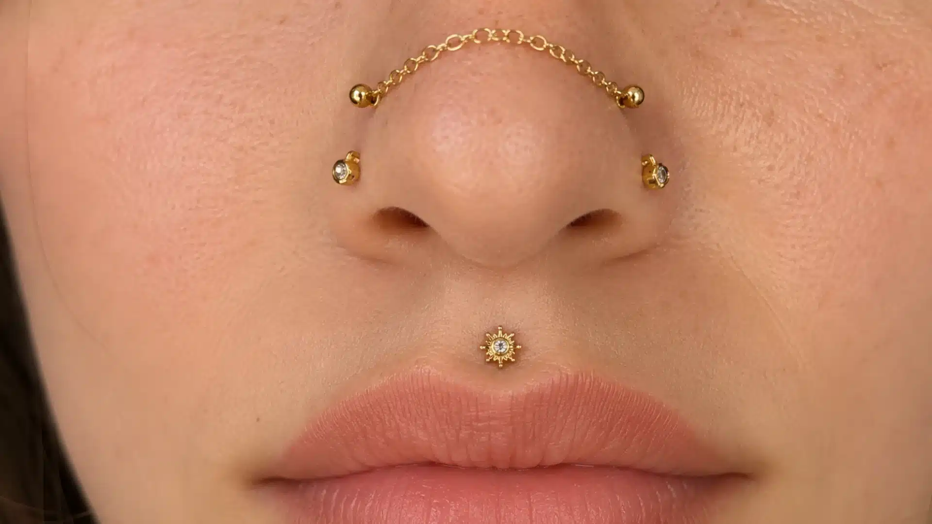 Close-up of gold nose piercings with a chain across the bridge and a sun-shaped medusa lip piercing on a person