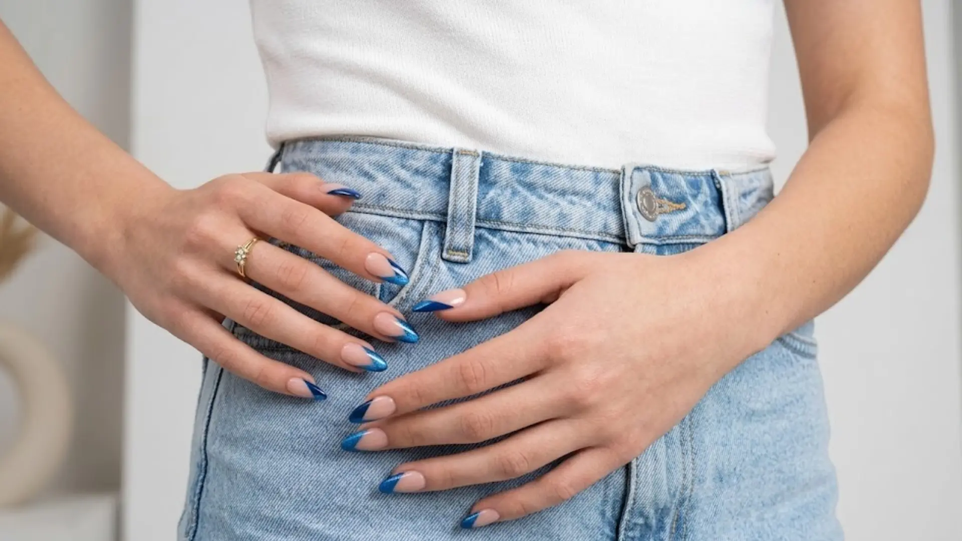 Close-up of hands with geometric blue French tip nails, gold rings, white top, and light blue denim jeans
