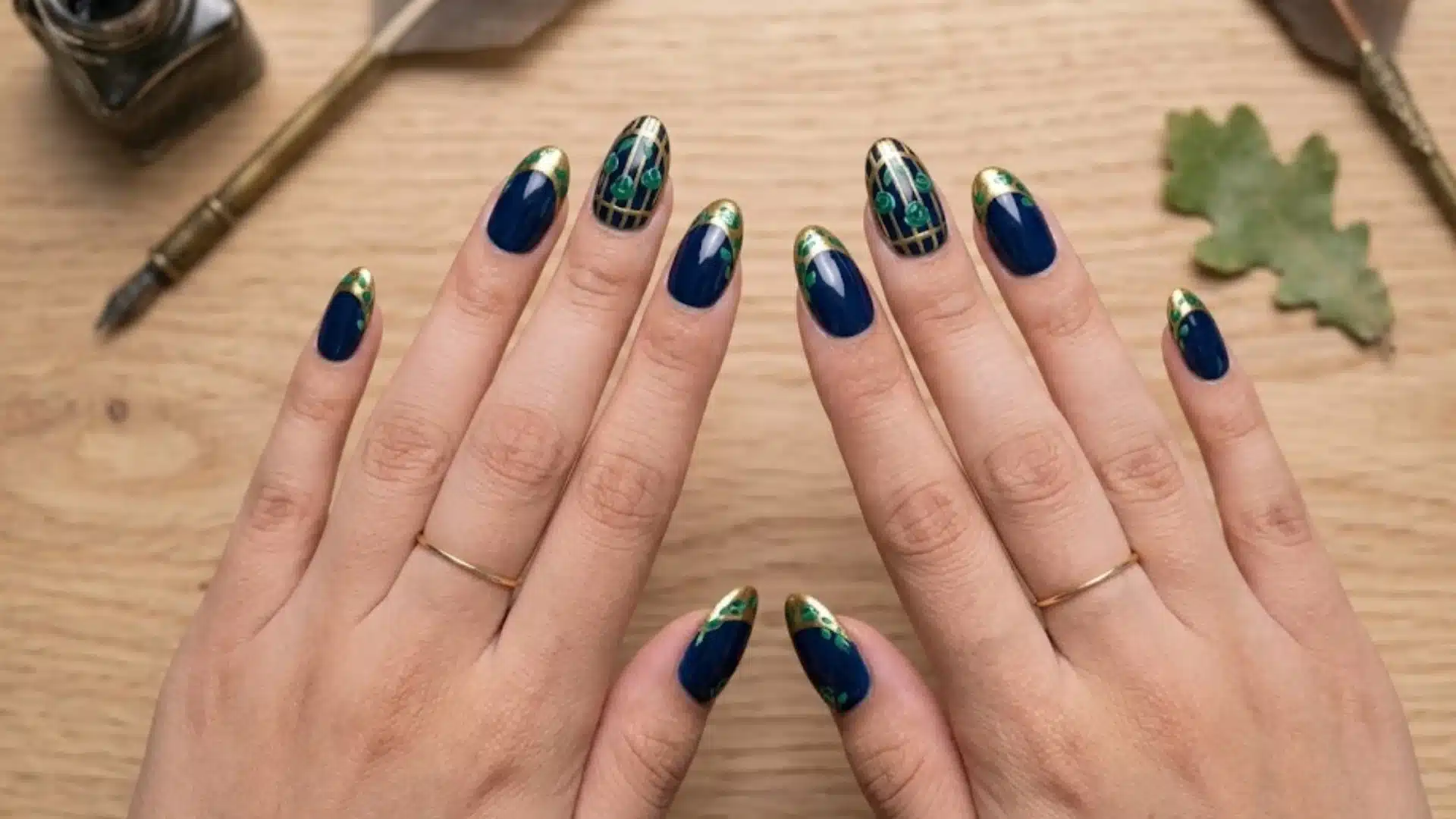 Dark blue nails with gold tips and intricate green accents, hands placed on wooden table