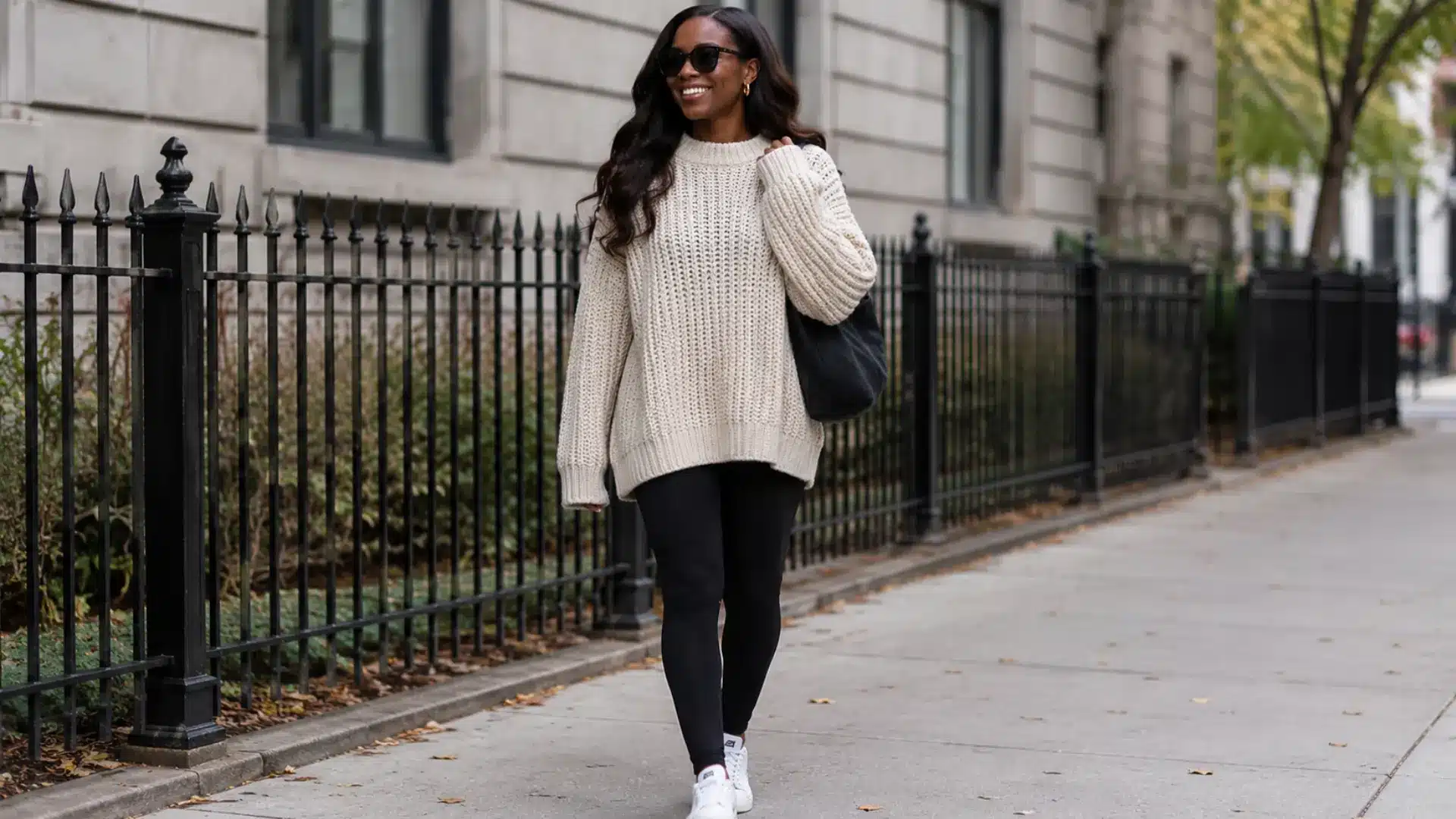 Dark skinned woman in oversized chunky sweater and black leggings walking on a city sidewalk