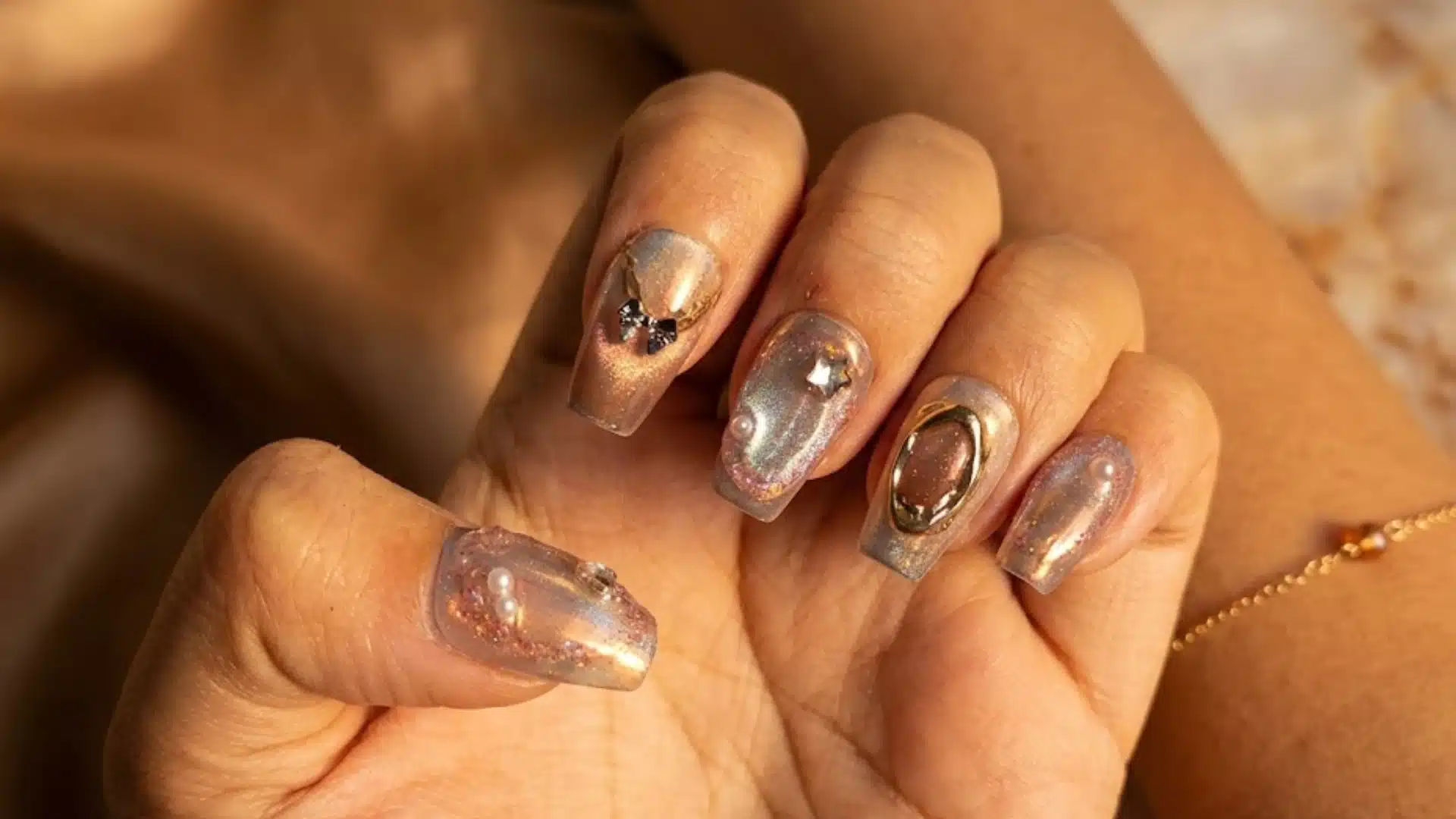 Glossy nude nails with gold chrome accents, pearls, and star details in warm golden sunlight