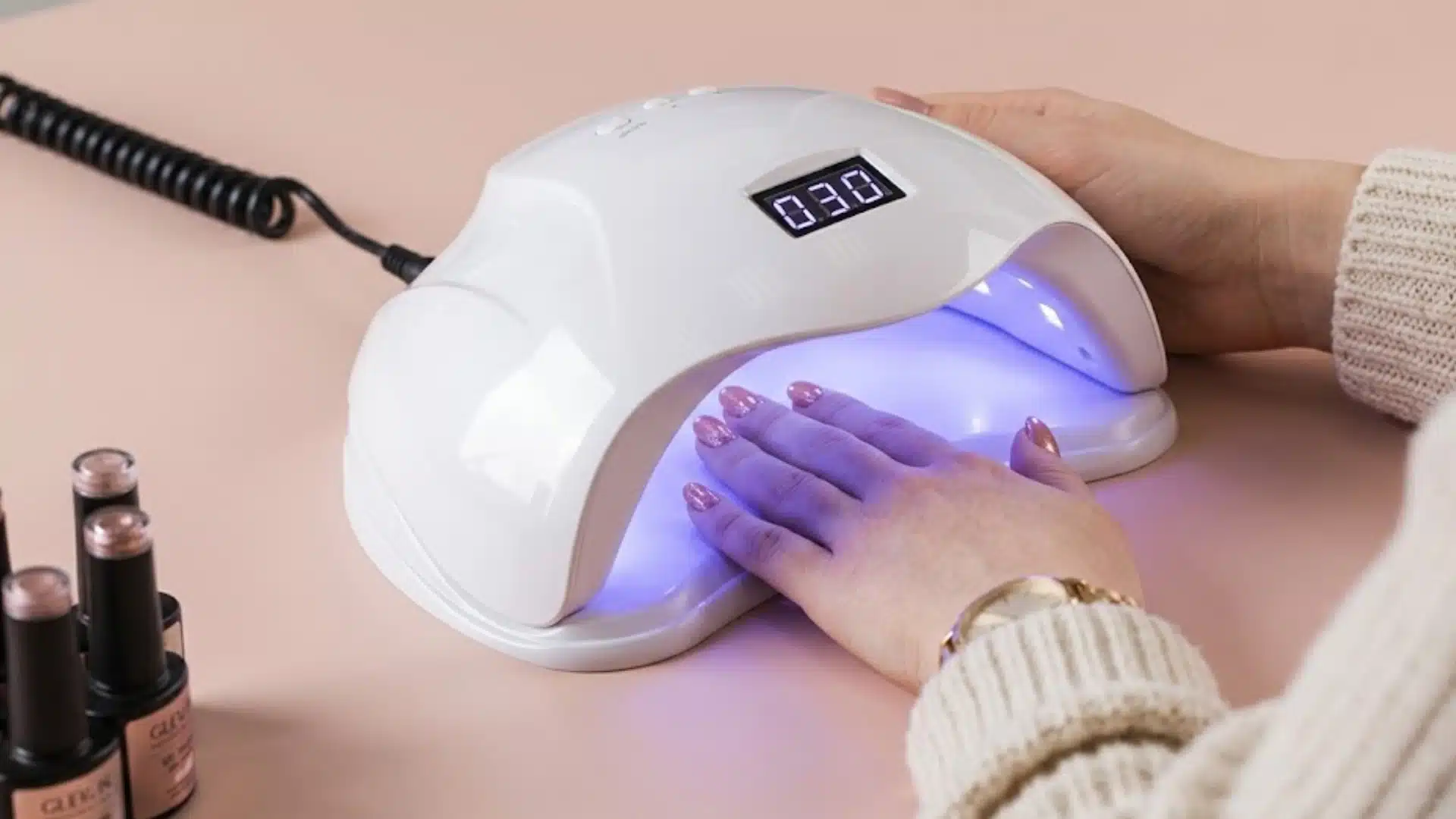 Hand curing pink gel nails under a UV nail lamp with polish bottles on a salon table