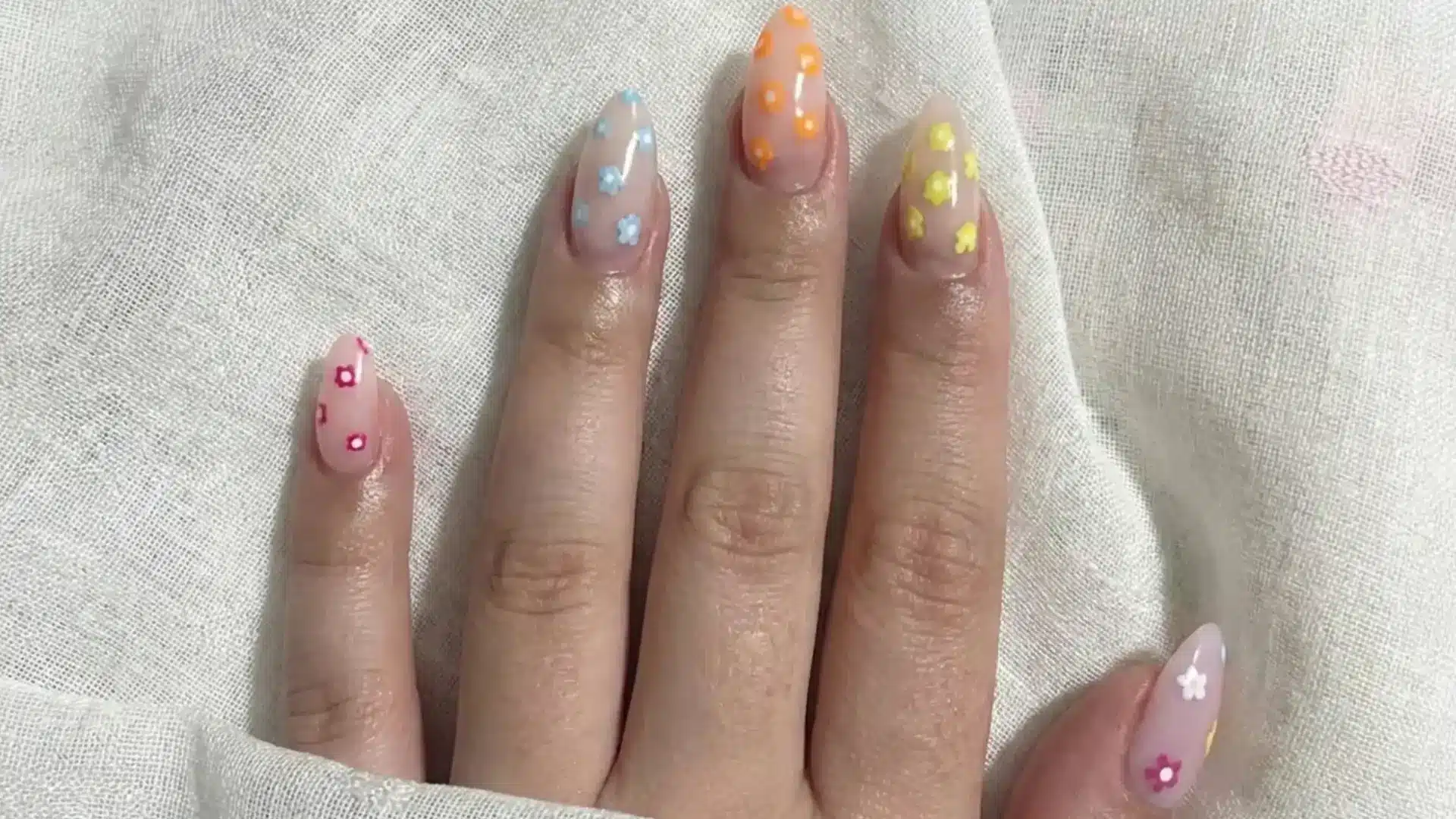 Hand with almond nails featuring pastel floral designs in pink blue yellow on sheer nude base