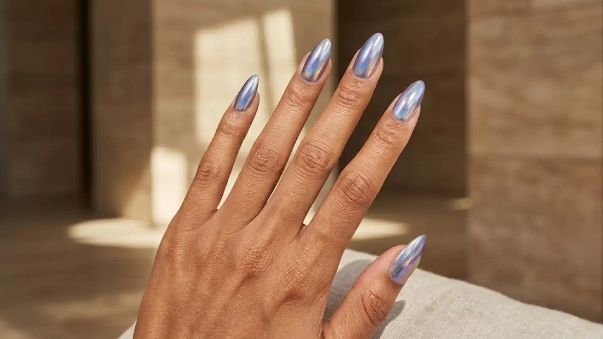 Hand with almond-shaped chrome nails in soft iridescent finish, glossy metallic shine in warm lighting