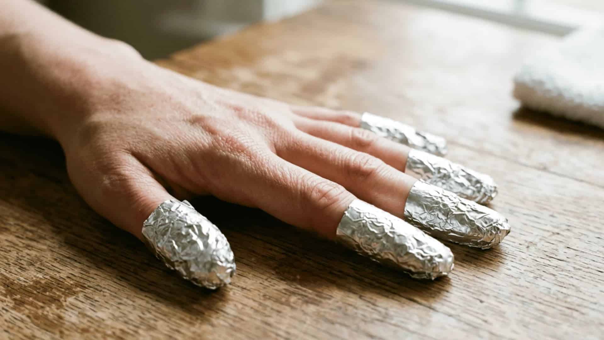 Hand with foil wrapped fingertips during gel nail removal on a wooden table