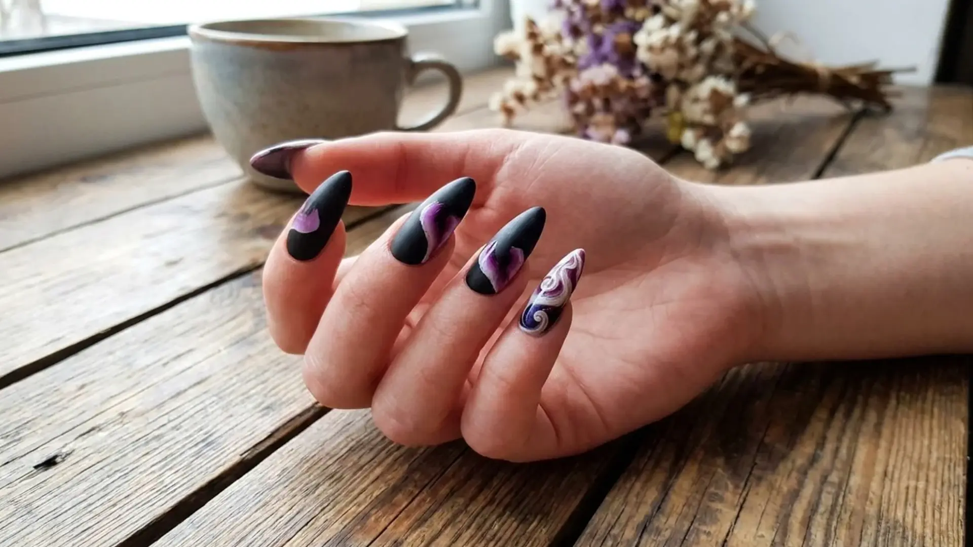 Hand with matte black almond nails featuring purple swirl nail art resting on wooden table near coffee cup