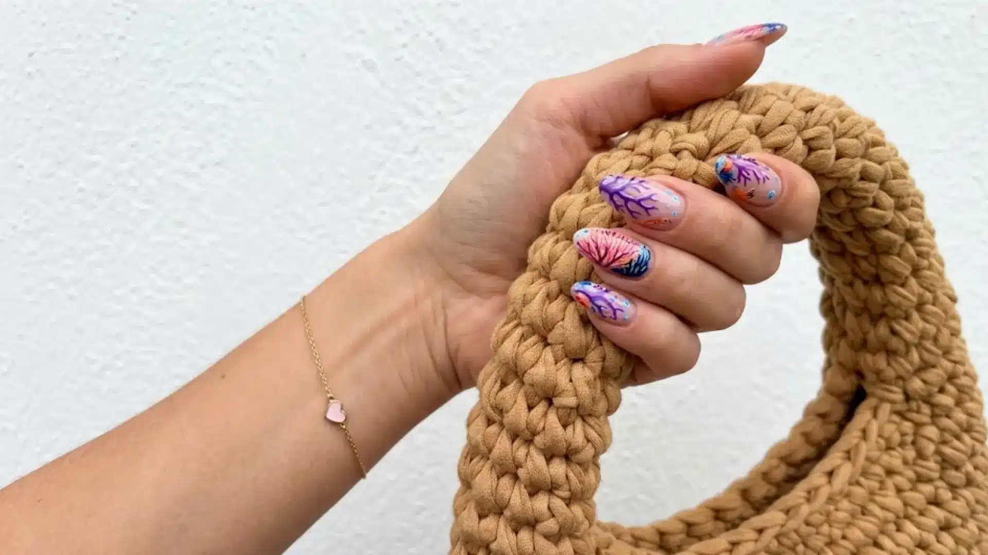 Hand holding a beige knitted bag with colorful nail art featuring shells and coral reef designs wearing delicate bracelet