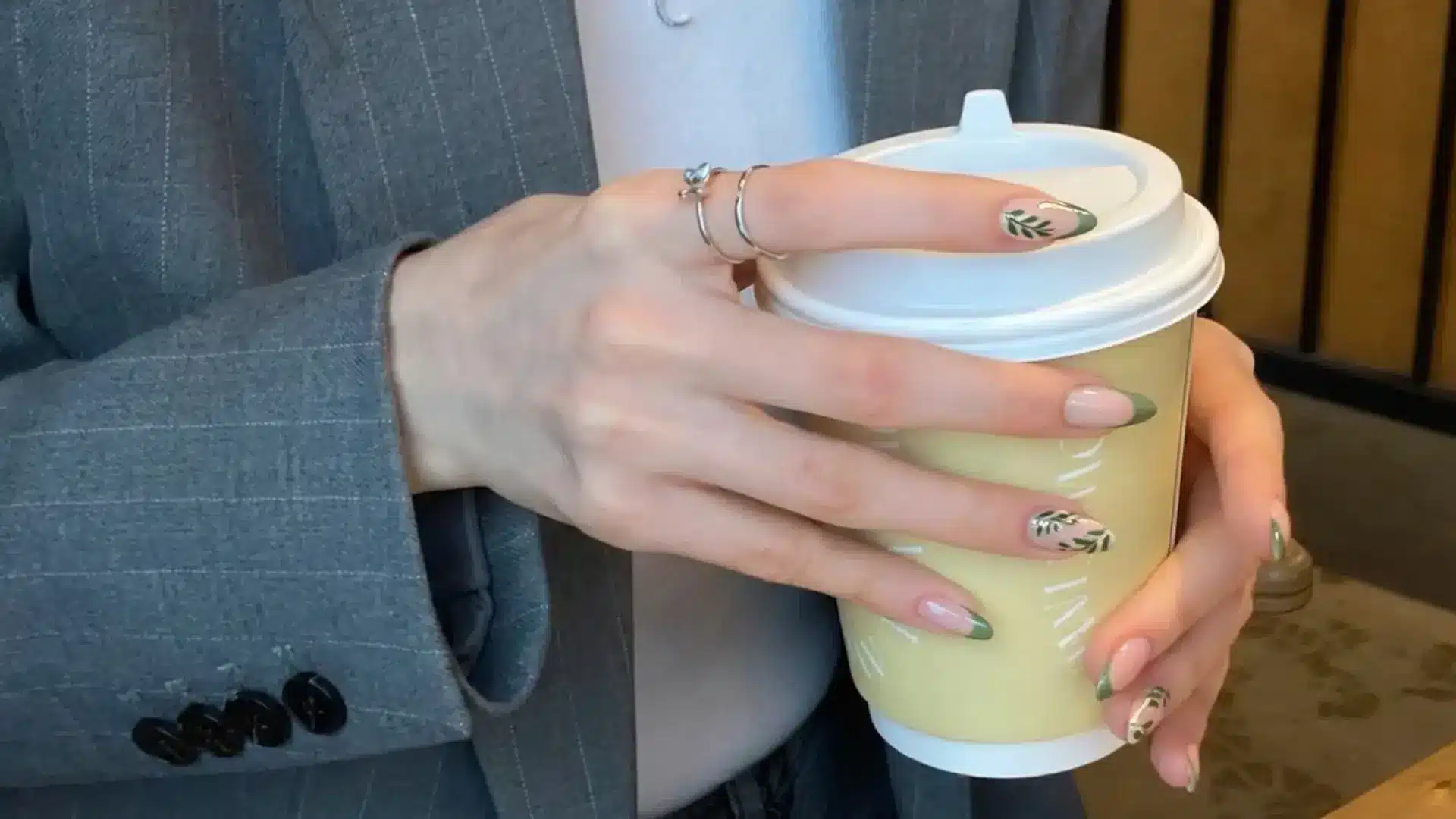 Hand holding coffee cup with sage green nail design featuring leaf art on almond shaped nude base nails