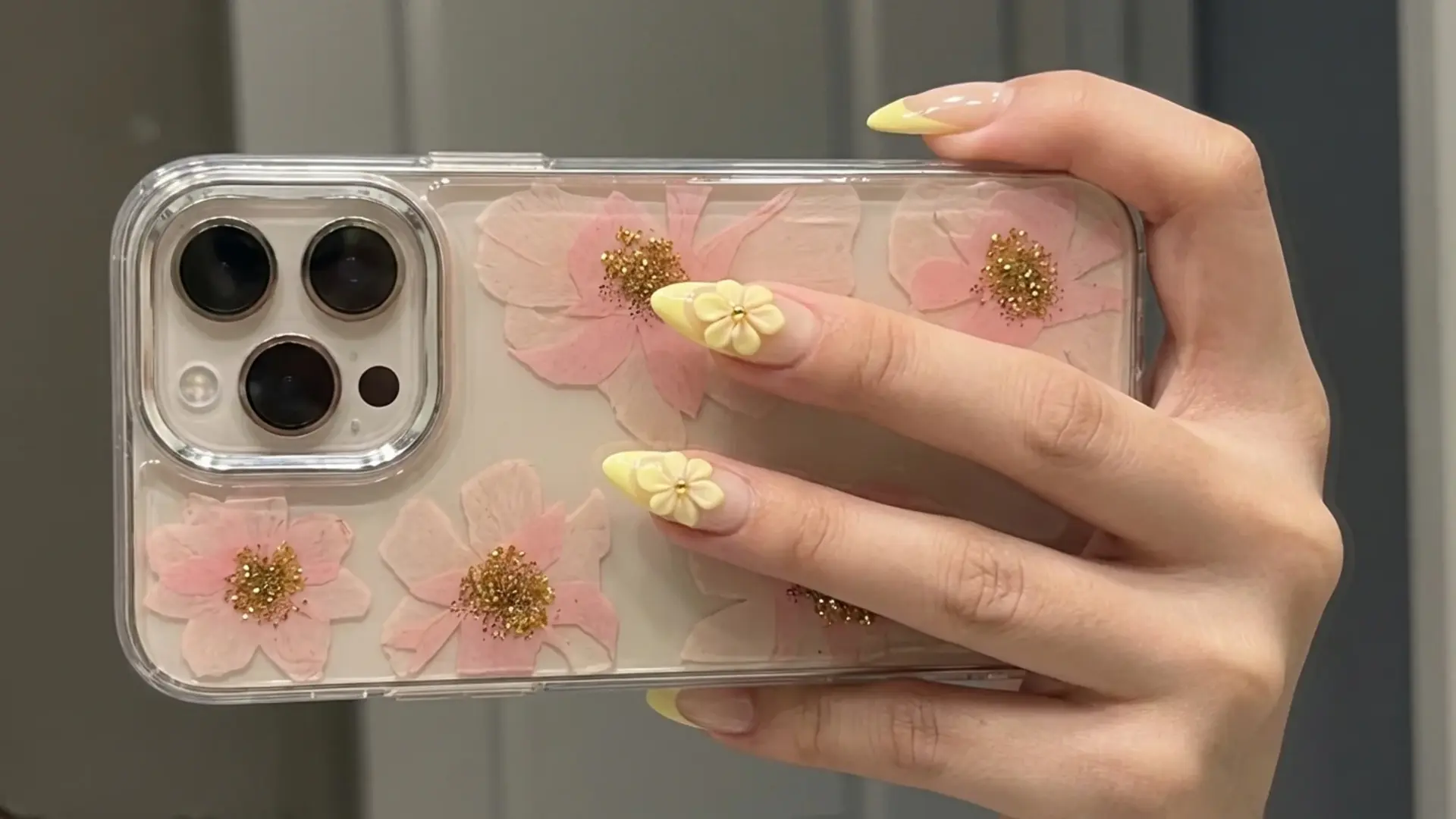 Hand holding phone with butter yellow nails featuring soft pastel yellow polish and small floral nail art accents