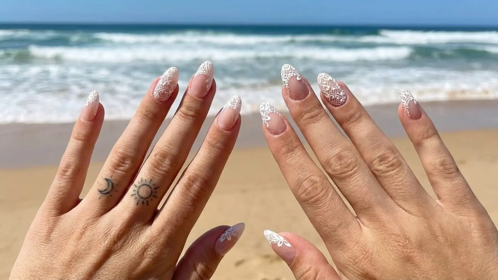 Hands with intricate white lace manicure held against beach waves showing bridemaids nail design