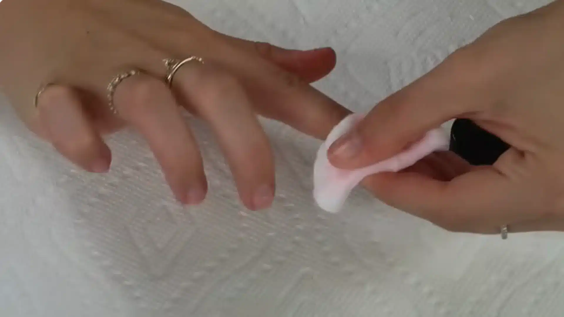 Hands with rings gently wiping fingernails using a cotton pad on a white textured surface