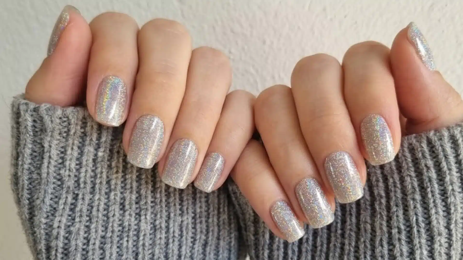 Hands with short square nails in holographic glitter polish, soft rainbow shine, glossy finish, cozy sweater background