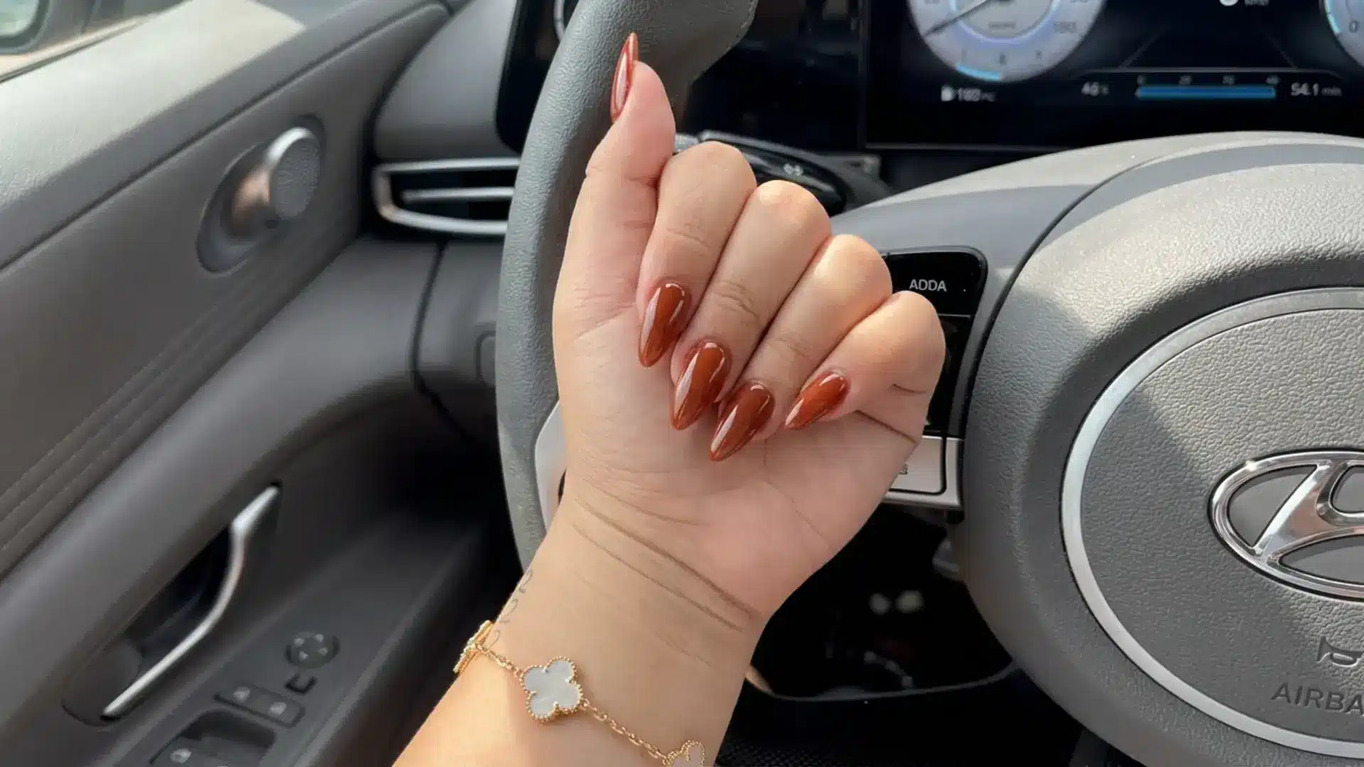 Hand showing terracotta nude almond nails with glossy warm brown polish inside car near steering wheel