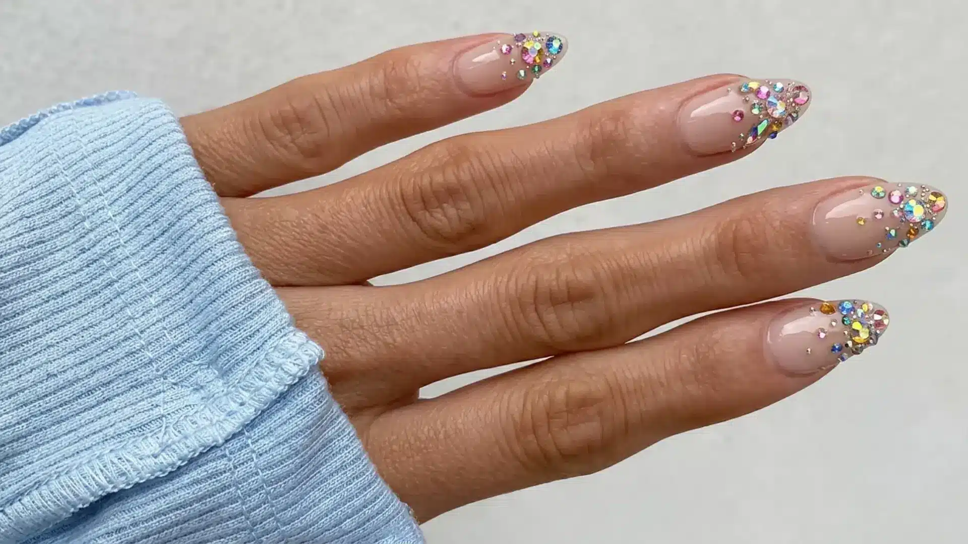 Hand with bejeweled crystal cluster nails featuring nude base and multicolor rhinestones concentrated at tips