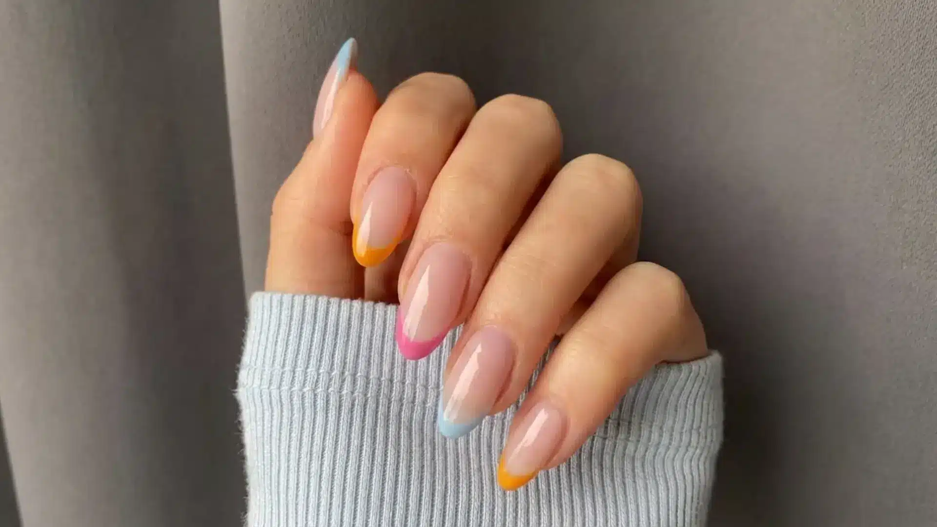 Hand with colored French tips featuring pastel blue, pink and yellow tips on nude almond nails against neutral background
