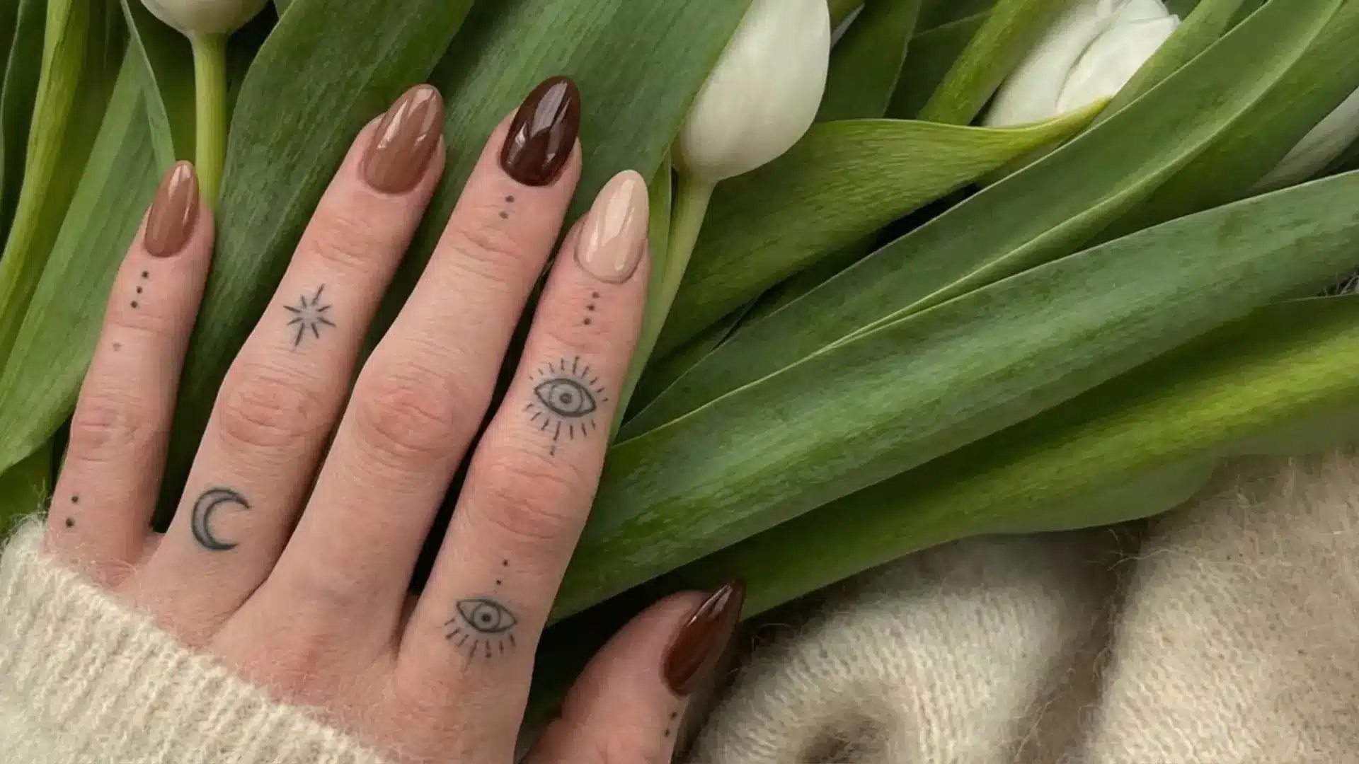 Hand with glossy brown and nude almond nails resting on green leaves with tulips showing minimal natural nail design