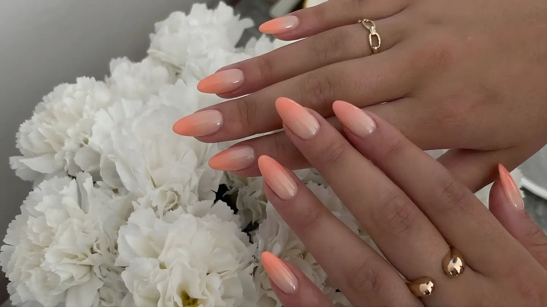 Hand with peach coral ombre nails showing soft gradient blend on long almond nails placed over white flowers