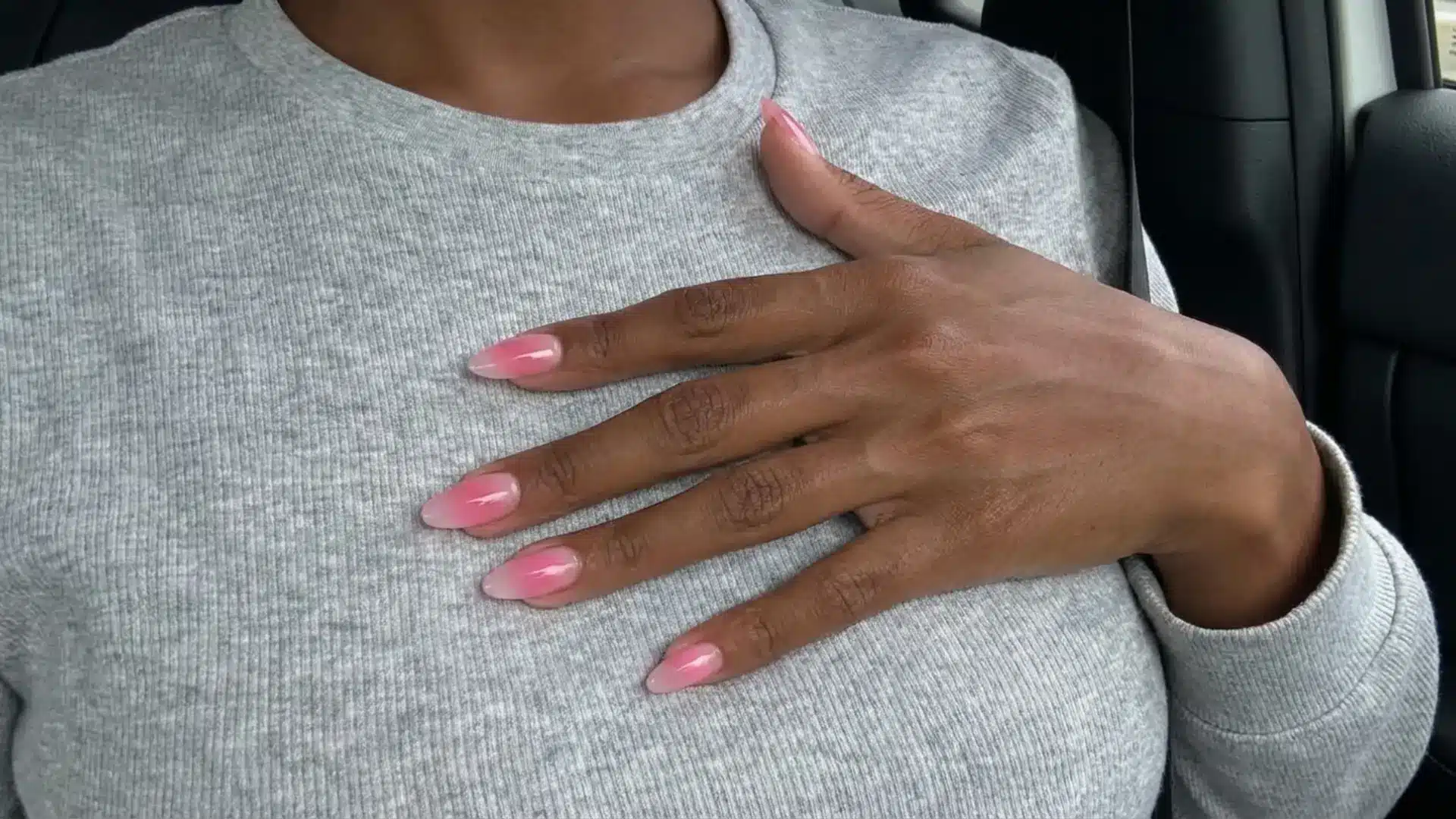 Hand with pink gradient aura nails resting on chest showing soft blended ombre effect on long almond shaped nails