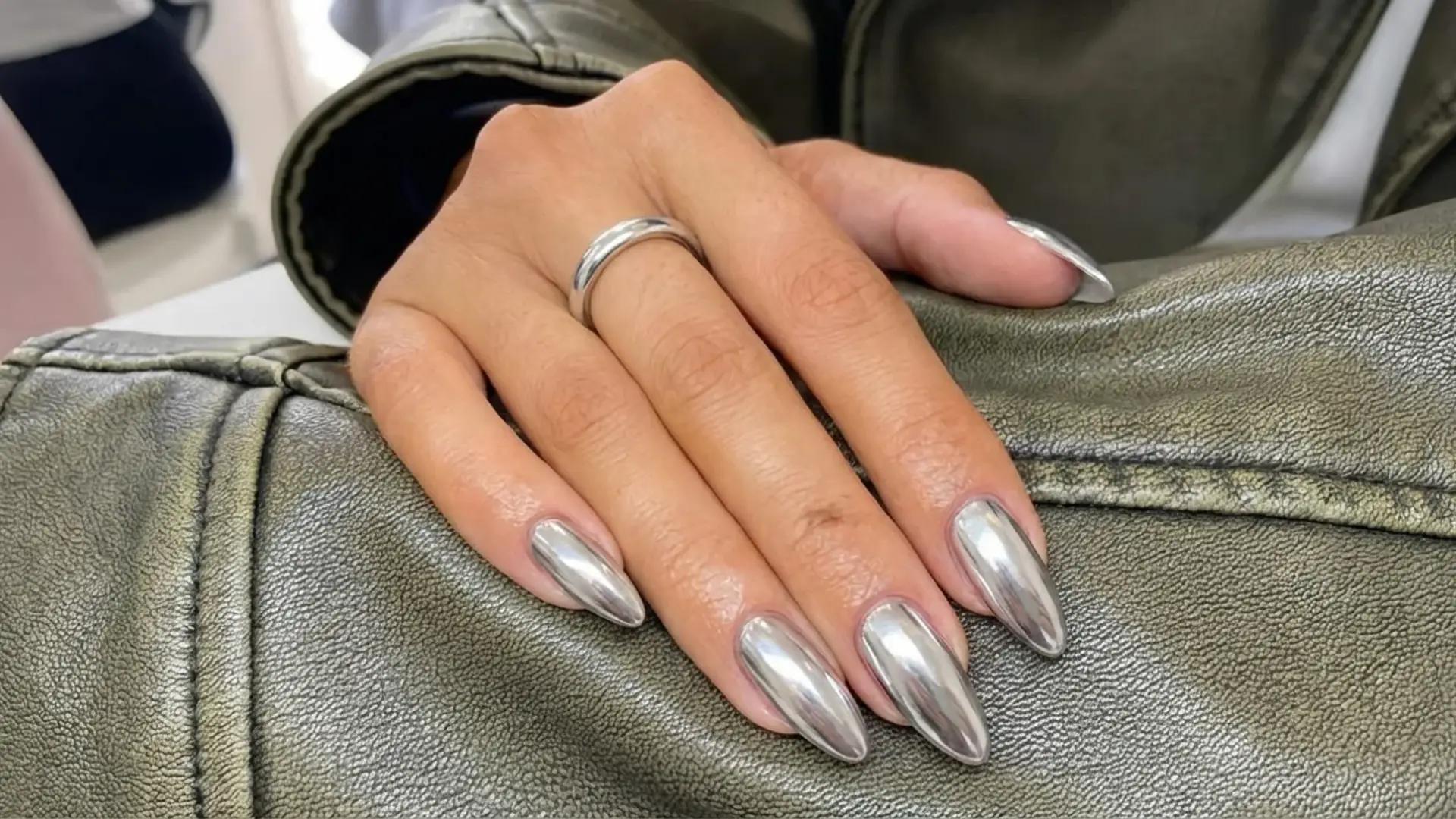 Hand with silver chrome almond nails showing reflective metallic finish resting on textured leather jacket