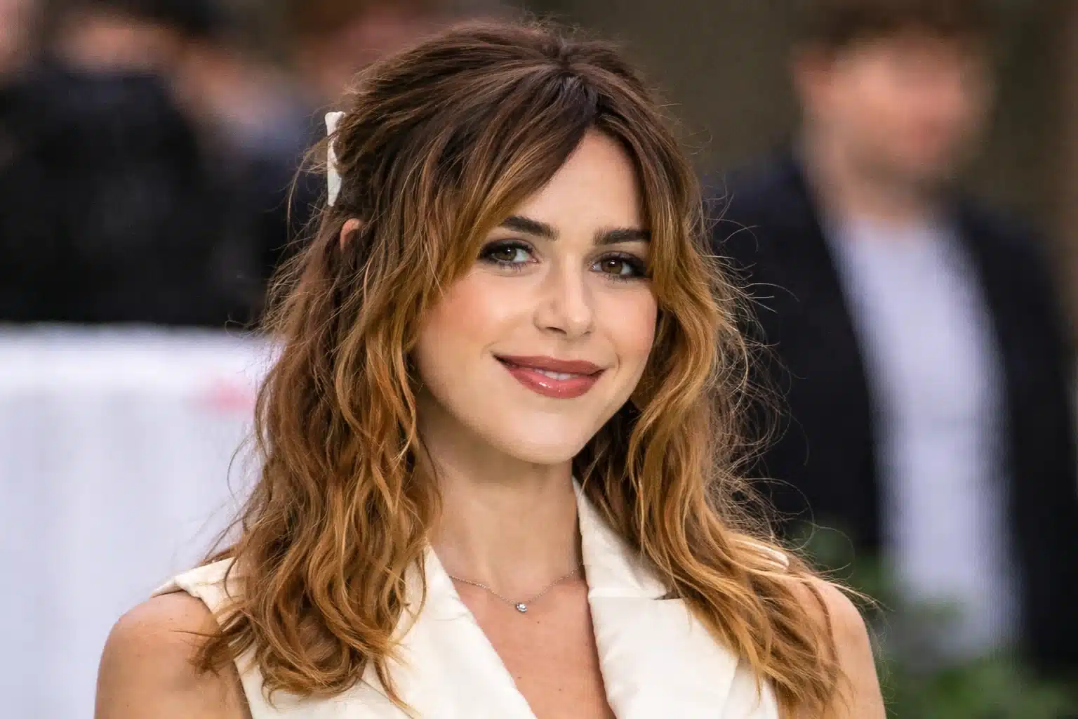 Kiernan Shipka with soft features and warm-toned makeup smiling outdoors, wearing a cream outfit with loose waves and face-framing bangs