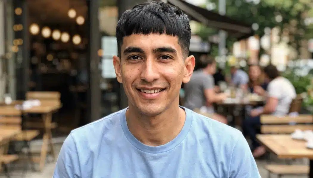 Man with a French crop haircut, blunt fringe, and faded sides sitting outside a busy restaurant cafe