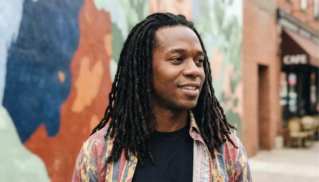 Man with shoulder-length locs standing outdoors near a colorful mural and brick storefronts