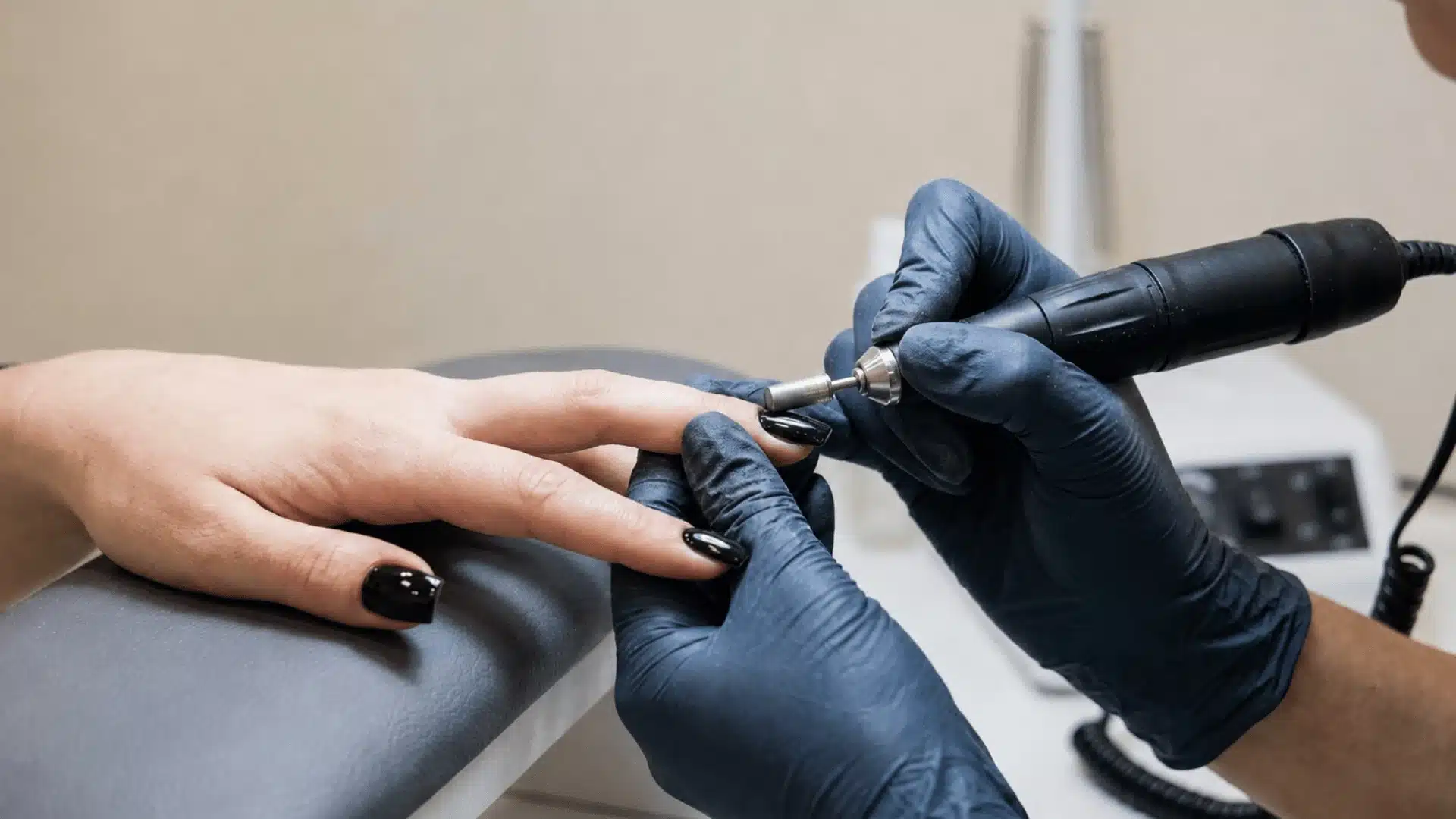 Nail technician wearing black gloves uses an electric nail drill on glossy black gel nails during a salon manicure