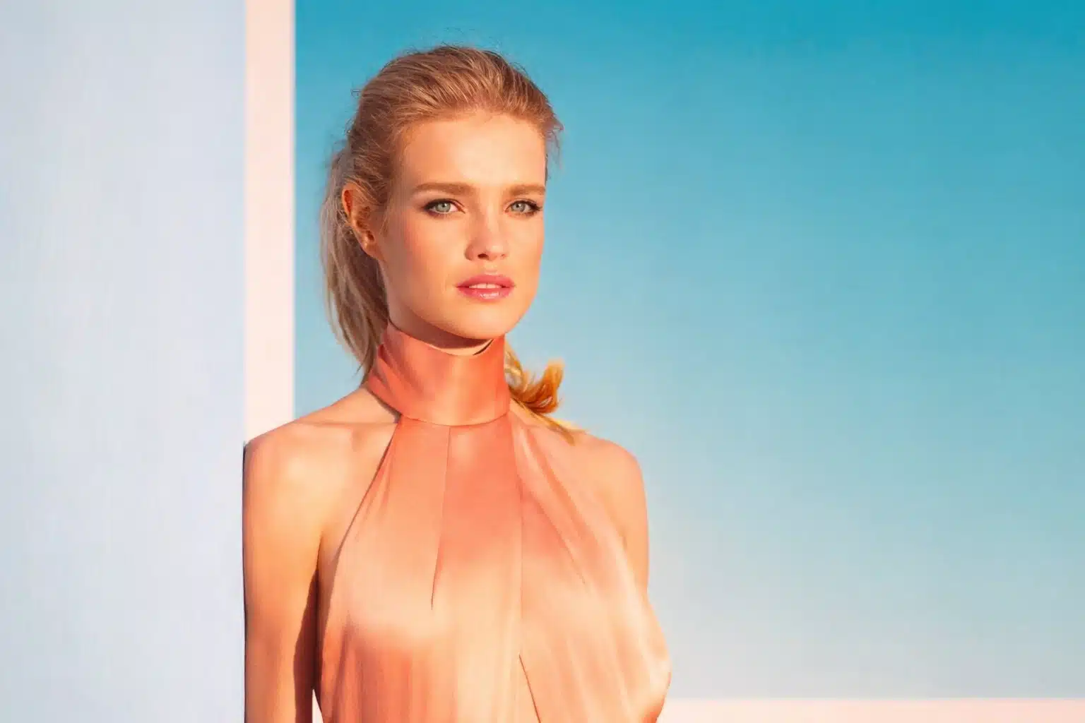 Natalia Vodianova with fair skin and blue eyes wearing a peach halter dress, softly lit by warm sunlight against a minimal blue background