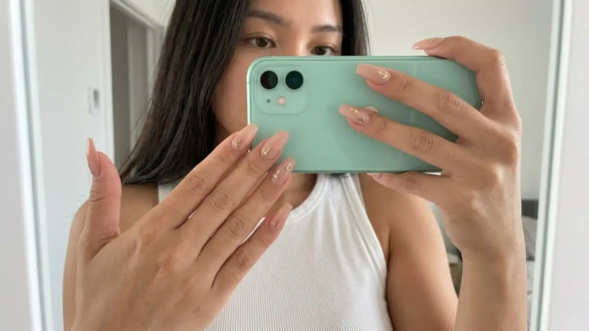 Nude almond nails with gold anchor accents held in selfie, minimalist manicure design for everyday chic style