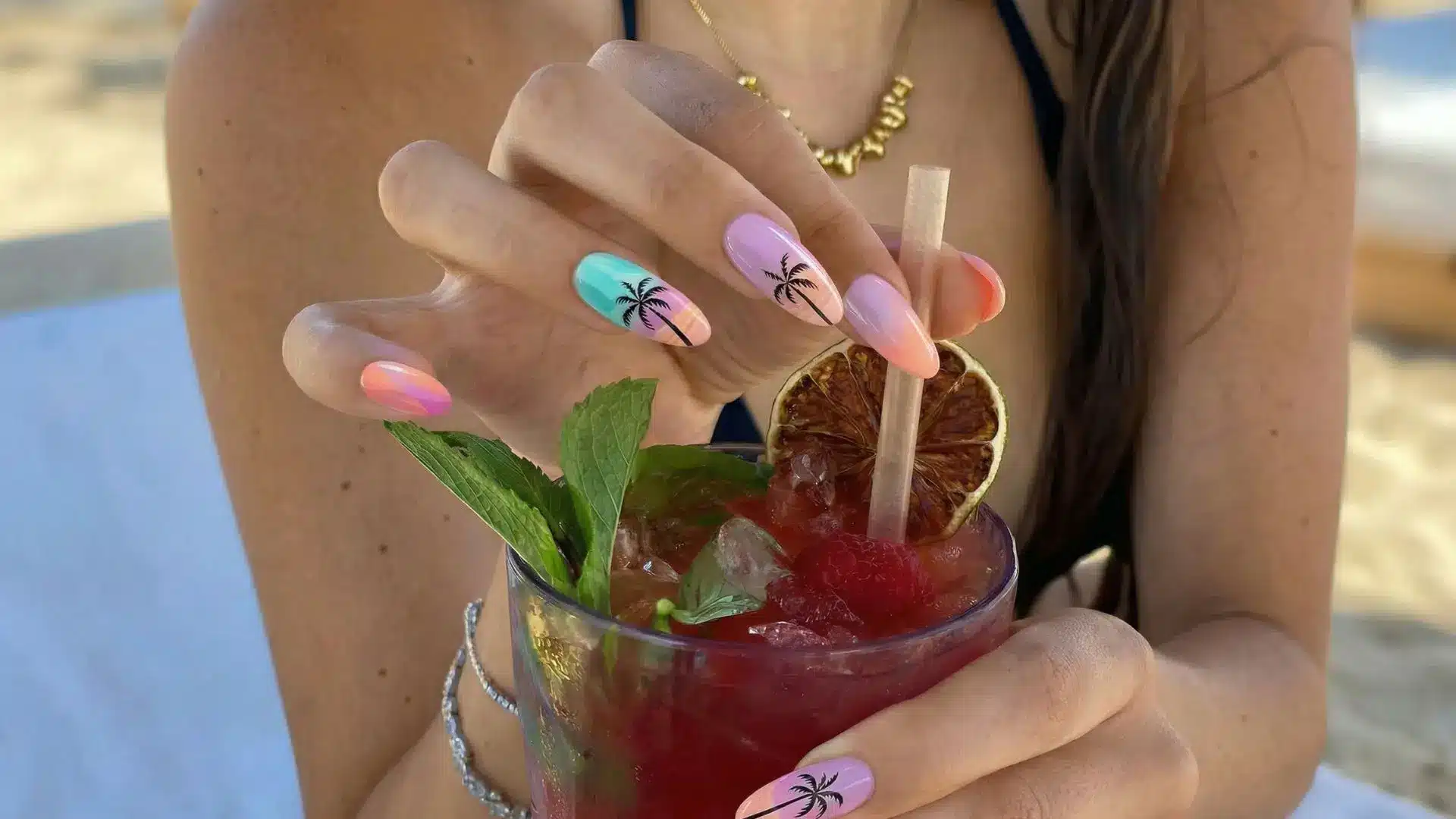 Pastel tropical sunset nails with palm tree designs holding a beach cocktail with mint garnish and citrus slice