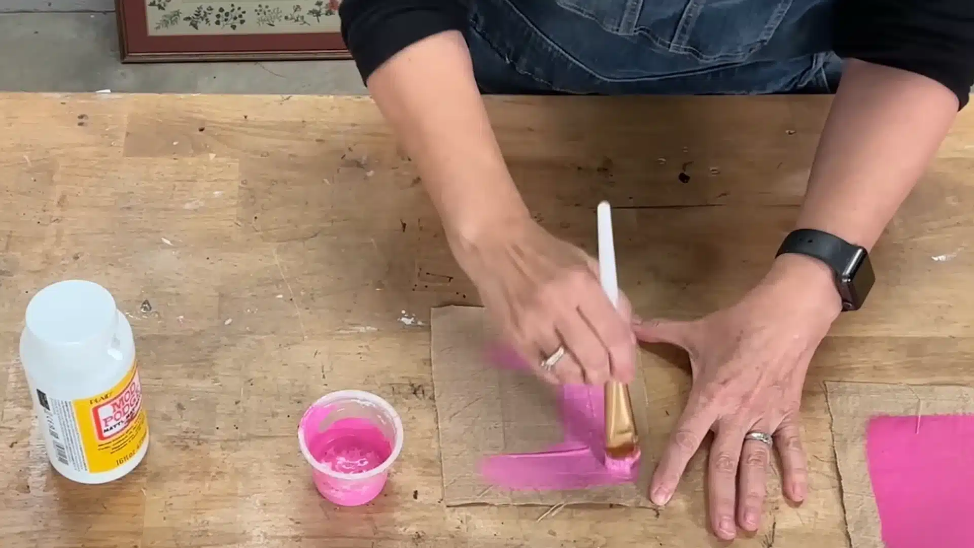 Person applying pink homemade fabric paint to a piece of fabric using a paintbrush on a wooden table