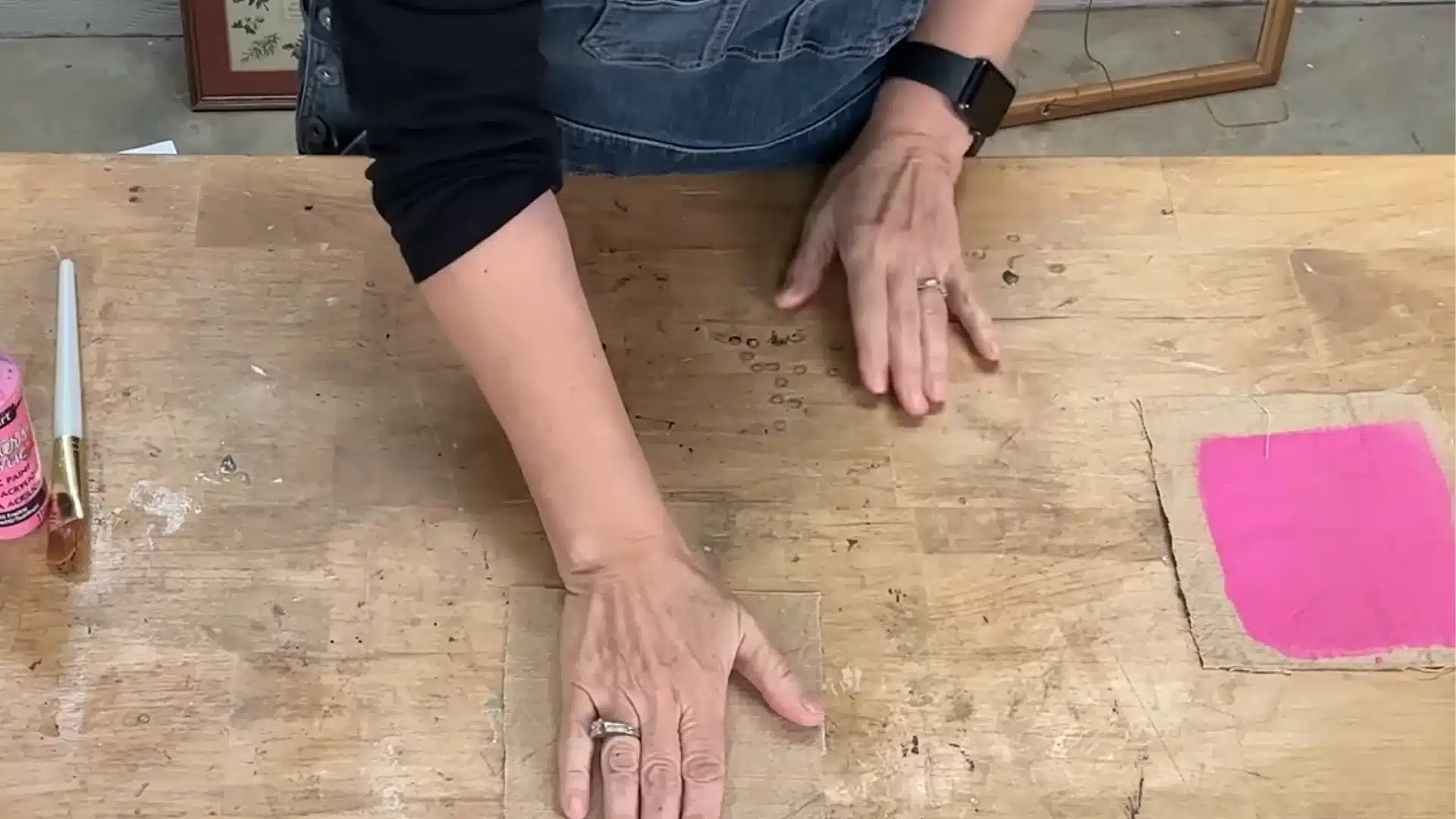 Person preparing wooden craft workspace with pink acrylic paint, brush, and painted fabric square