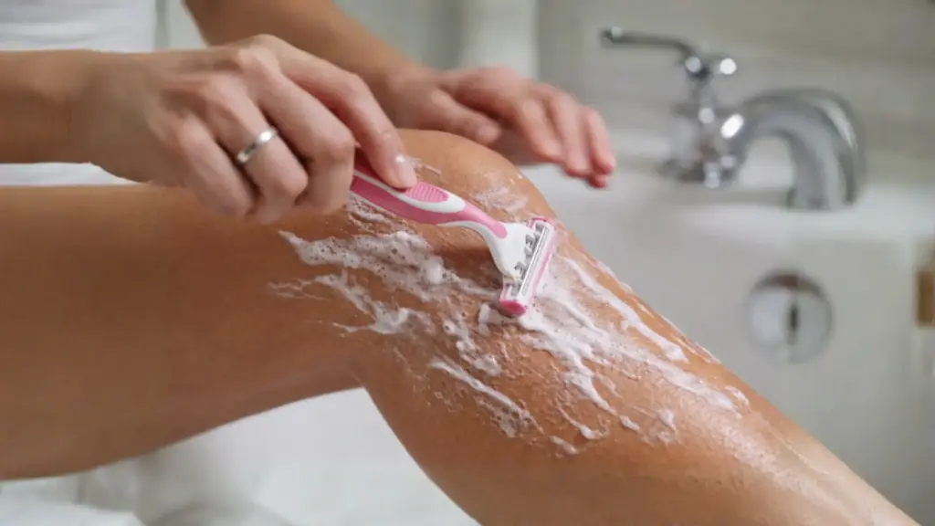 Person shaving leg with razor and shaving cream in bathroom showing hair removal step after spray tan care