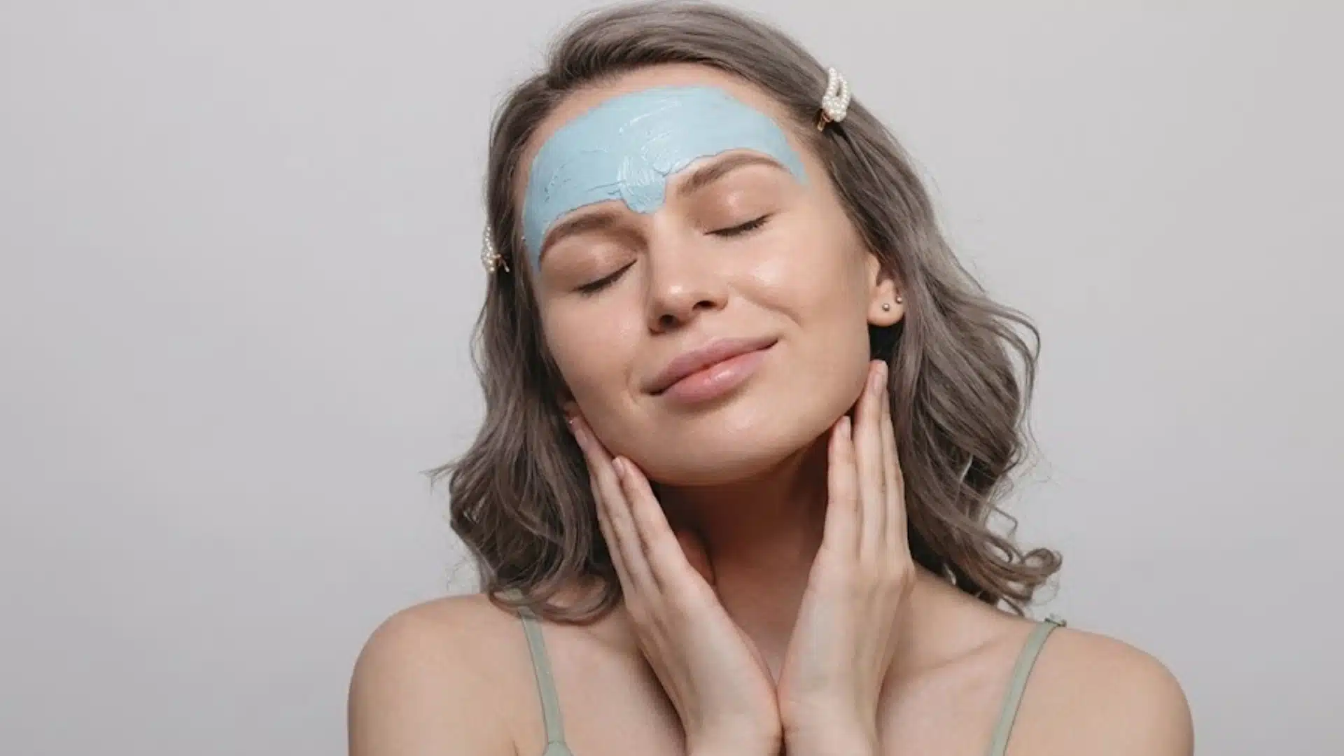 Person with blue clay mask applied on forehead, eyes closed, hands on face, showing skincare routine