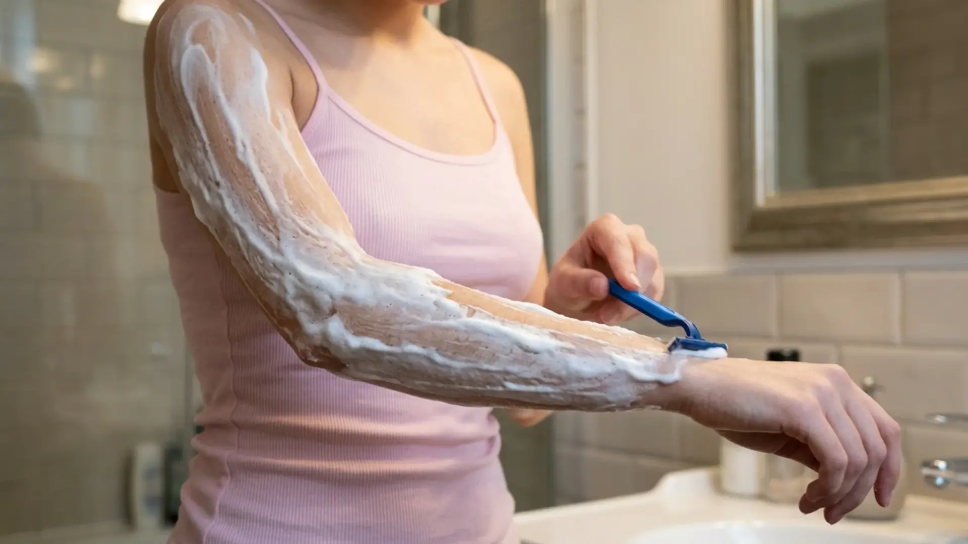 Person shaving arm with razor while applying thick shaving cream in bathroom near sink and mirror