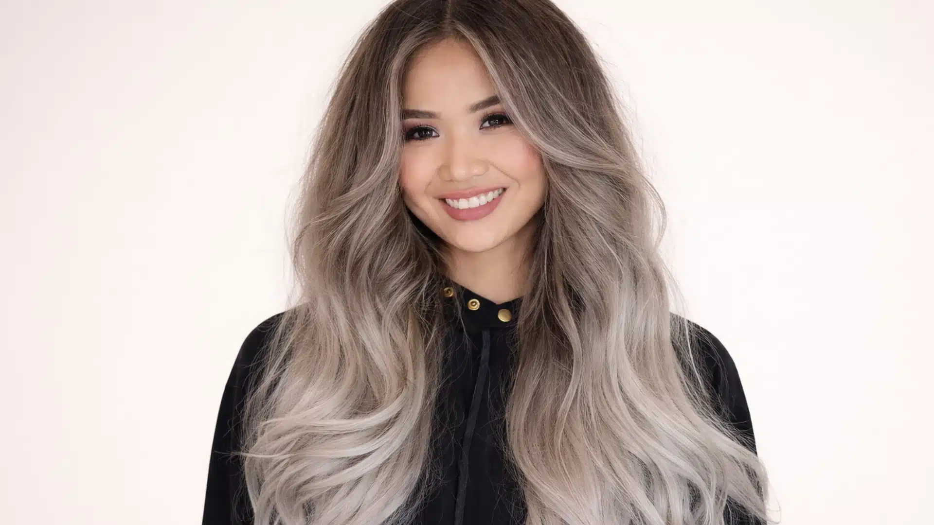 Portrait of a woman with long ash blonde balayage hair with soft waves and darker roots, wearing a black top against a clean solid beige background with focus on hair texture and color