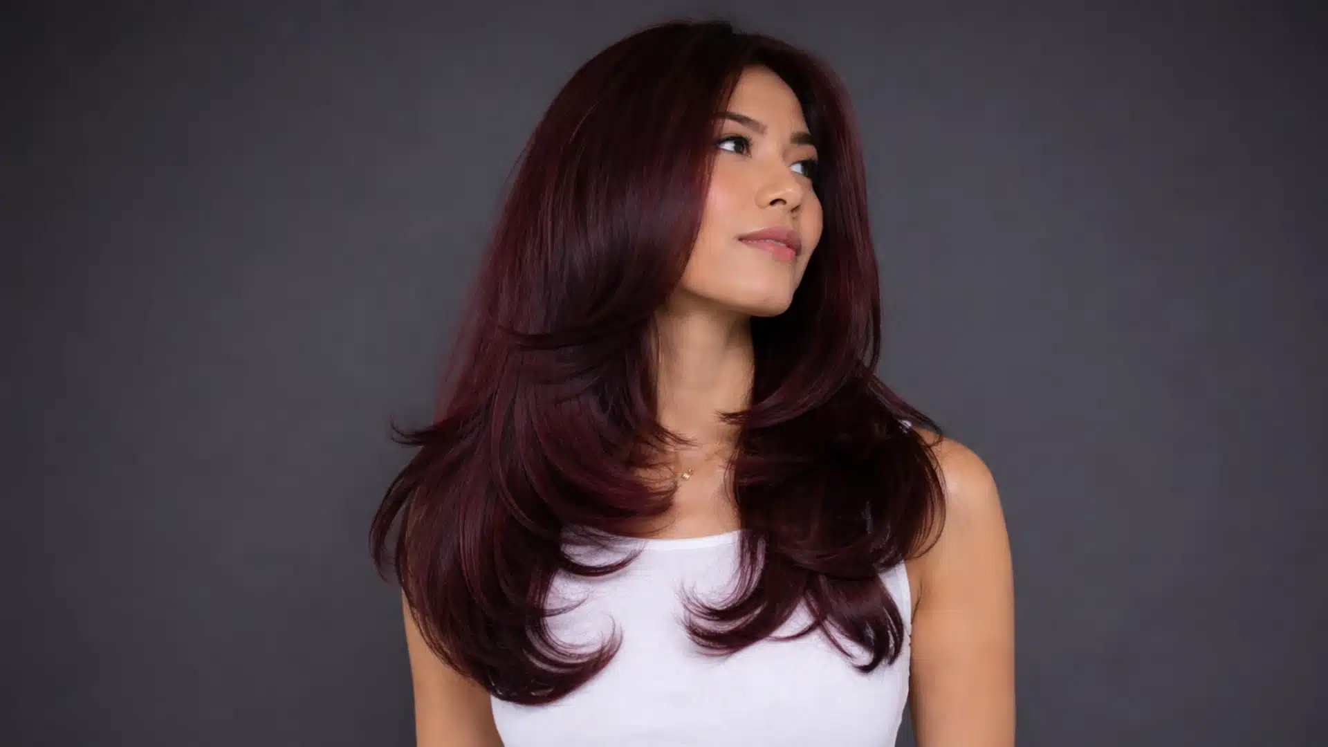 Portrait of a woman with long burgundy red layered hair, soft waves and shine, wearing a white top against a dark grey background with focus on hair color and texture